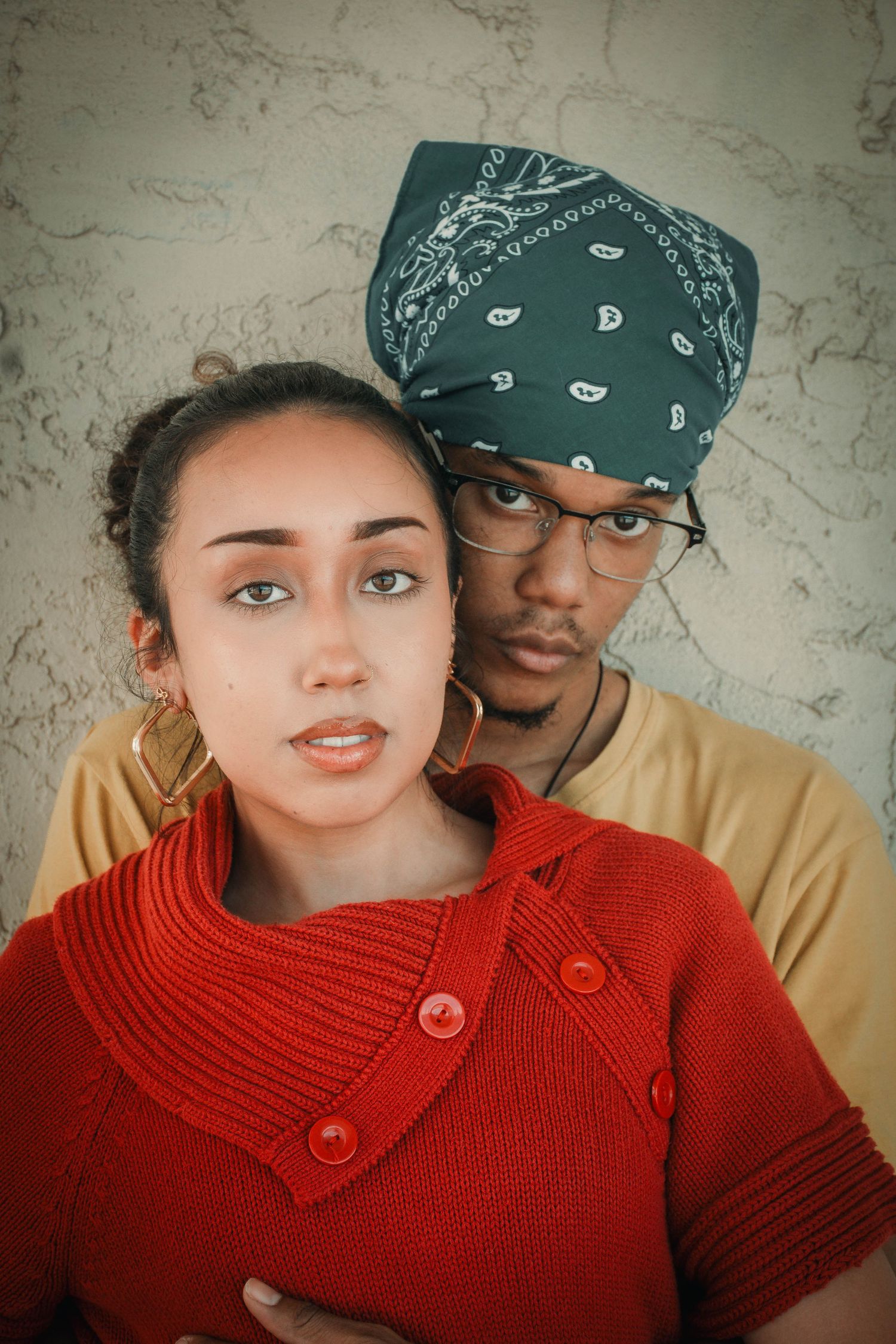 A young couple posing together, woman in red knit sweater, man wearing green paisley bandana and glasses.