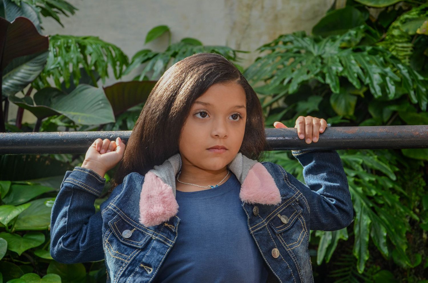 Young girl in denim jacket holding bar across shoulders in front of tropical green plants.