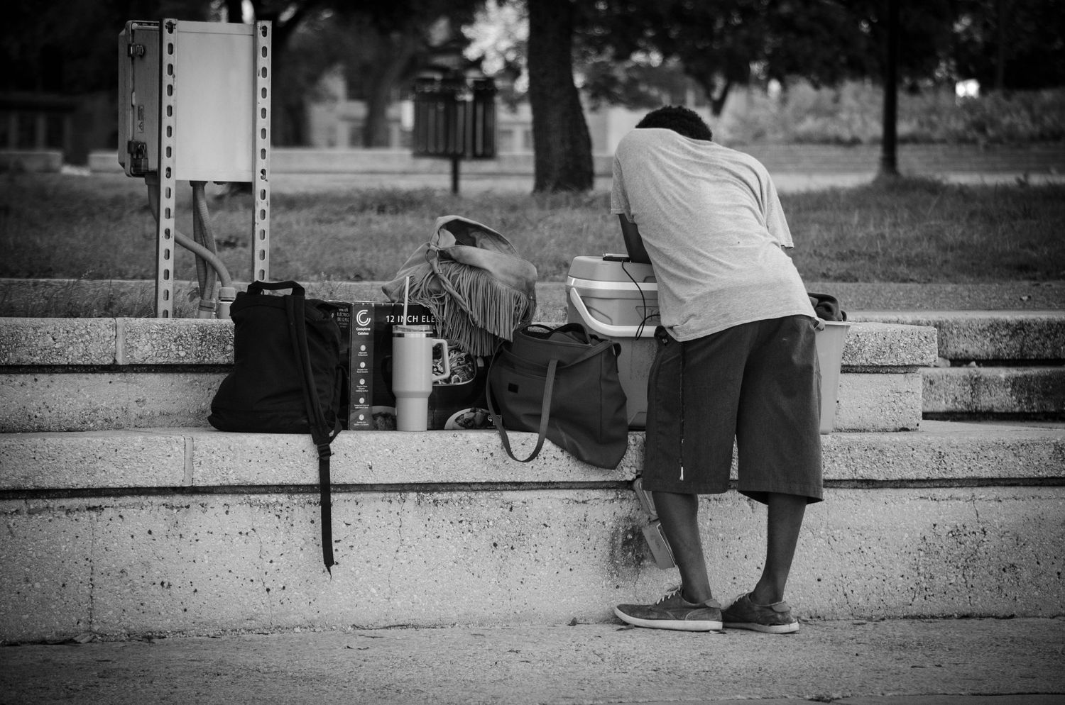 Black and white photo of a person leaning over a concrete ledge in a park with belongings nearby.