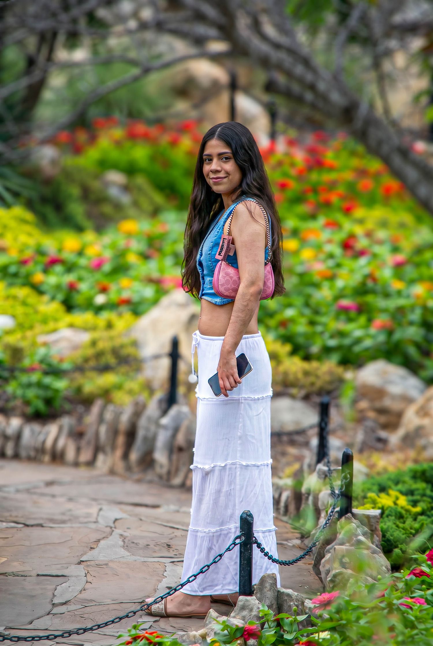 Young woman in colorful crop top and white maxi skirt posing on a stone garden pathway.