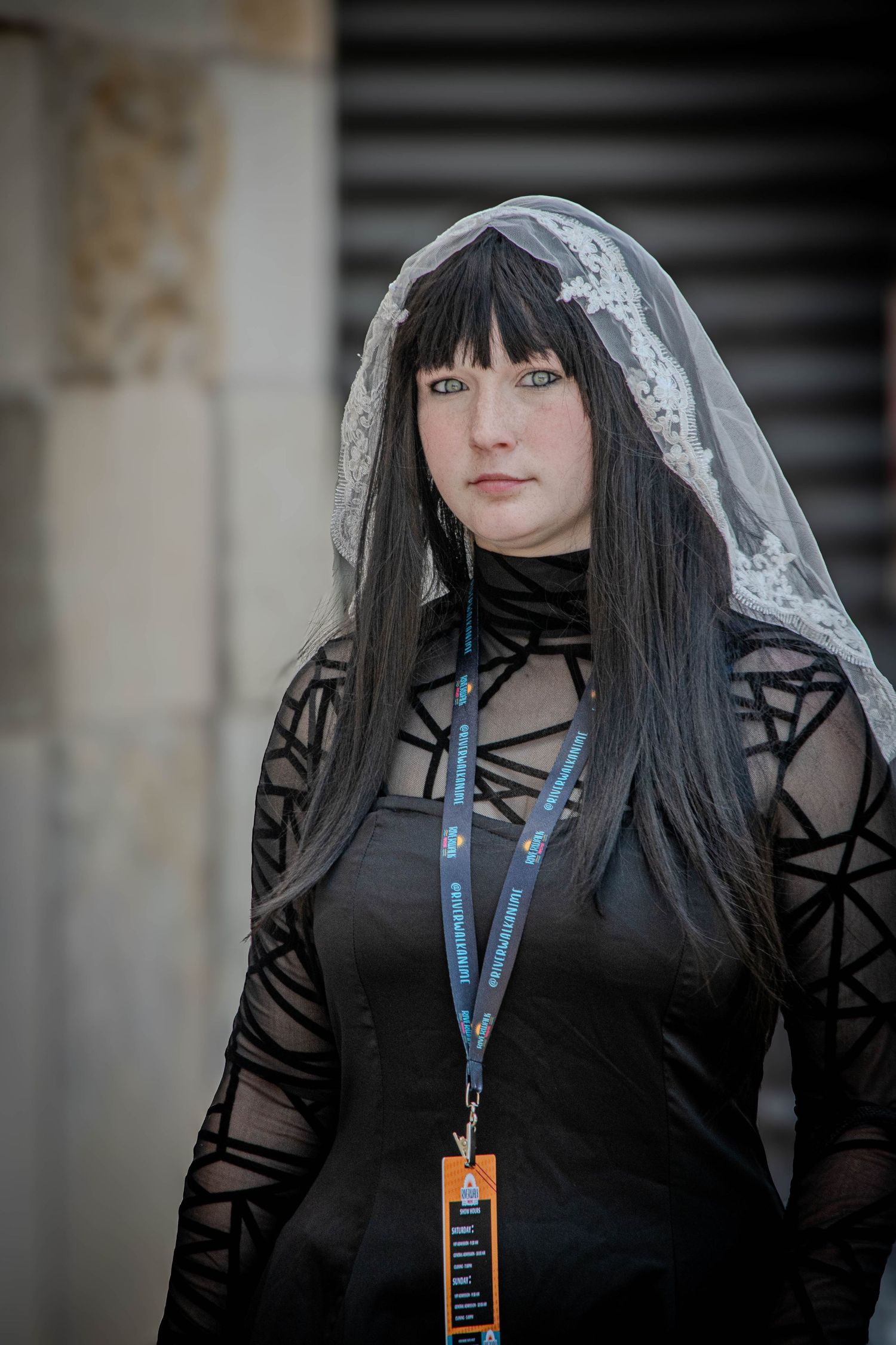 Woman in black gothic dress with lace veil and geometric sleeves wearing event lanyard.
