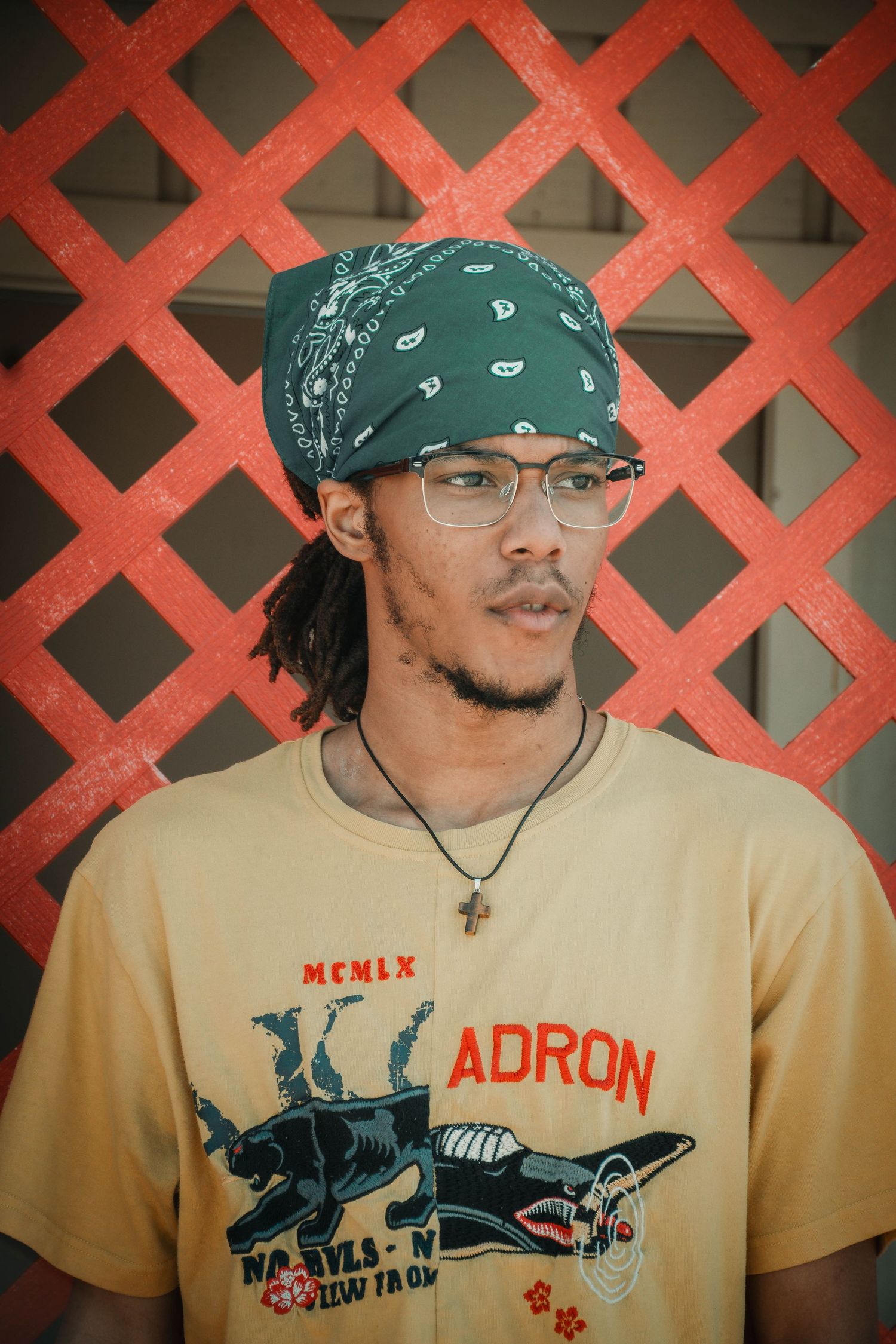 Young man wearing green bandana, glasses, and ADRON graphic tee poses against red lattice wall.