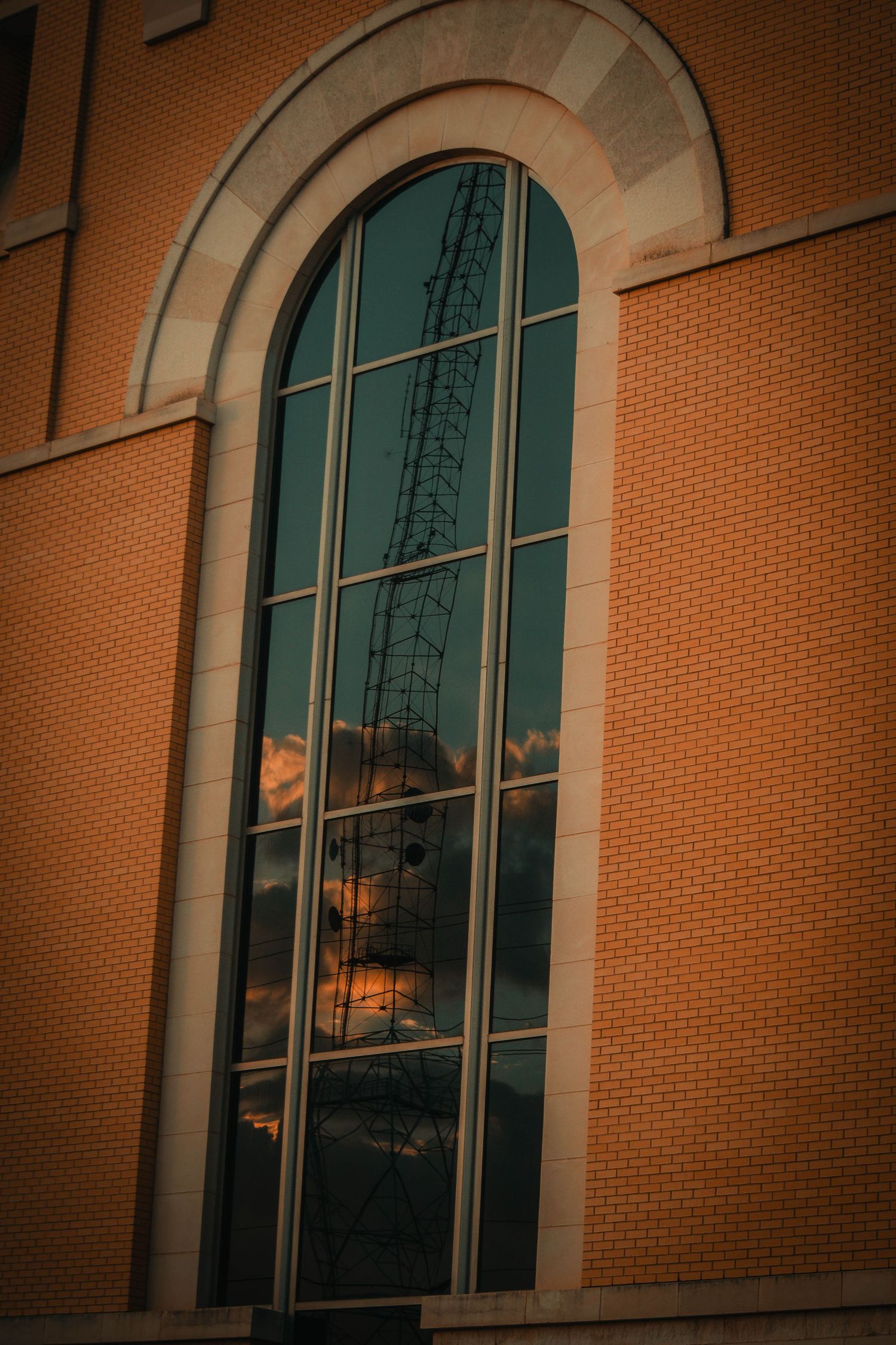 Arched glass window on brick building reflecting a tower crane and dramatic sunset clouds in warm golden light.