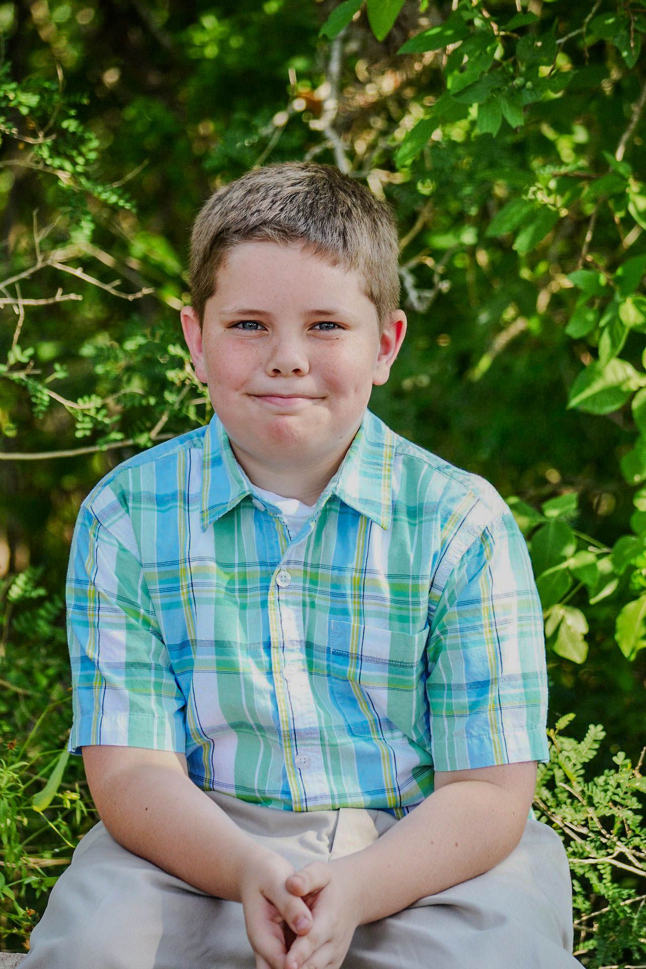 Smiling boy in plaid shirt sitting outdoors surrounded by green foliage.