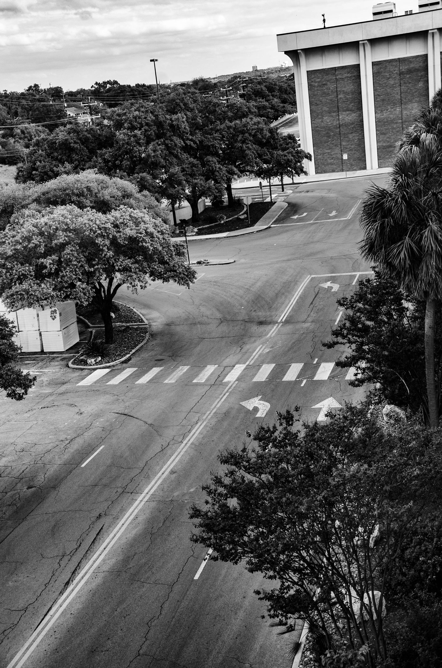 Aerial black and white view of an empty intersection with crosswalks and arrow markings near a large building.