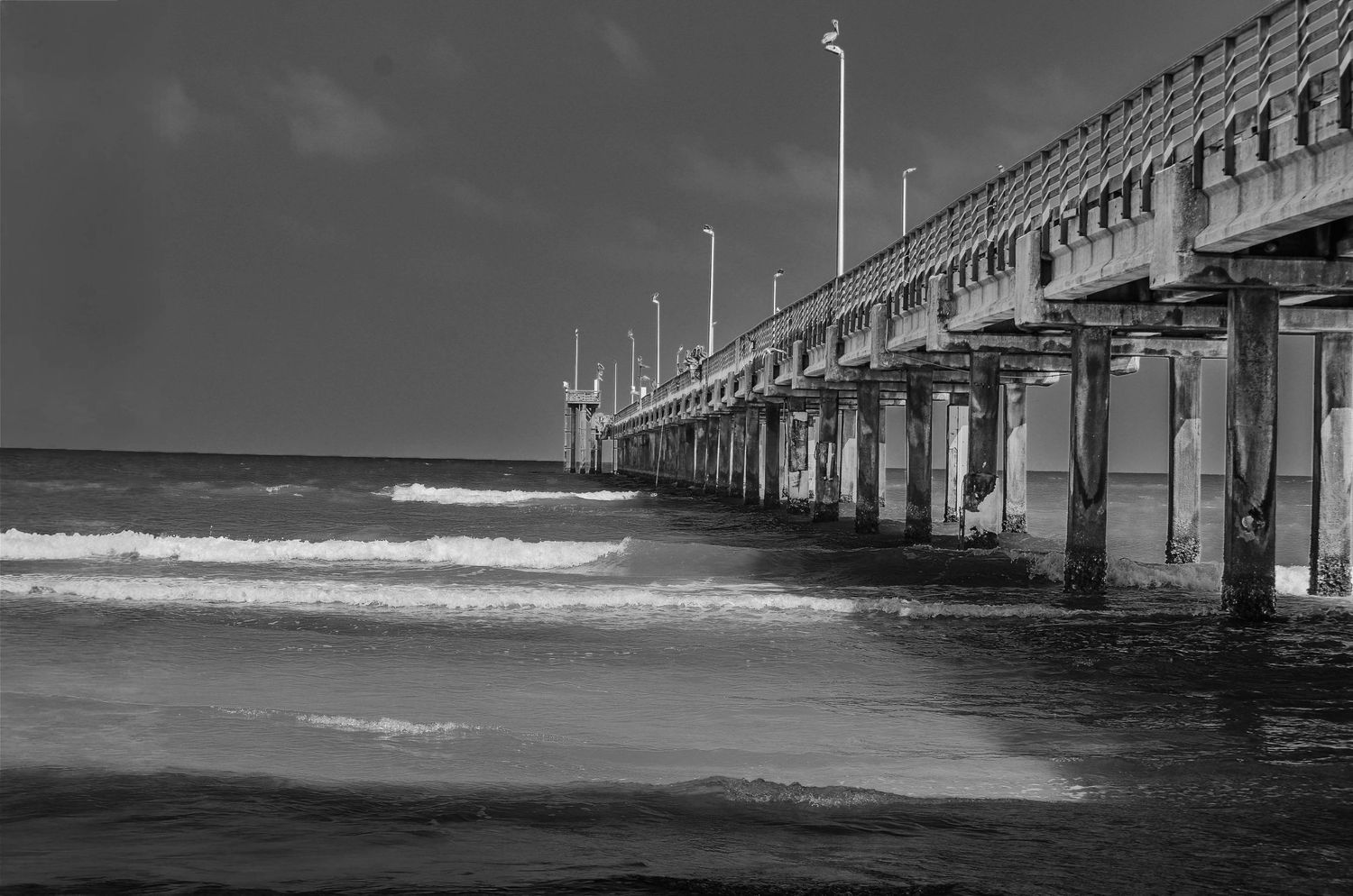 Black and white photo of a long ocean pier extending into the sea with crashing waves.