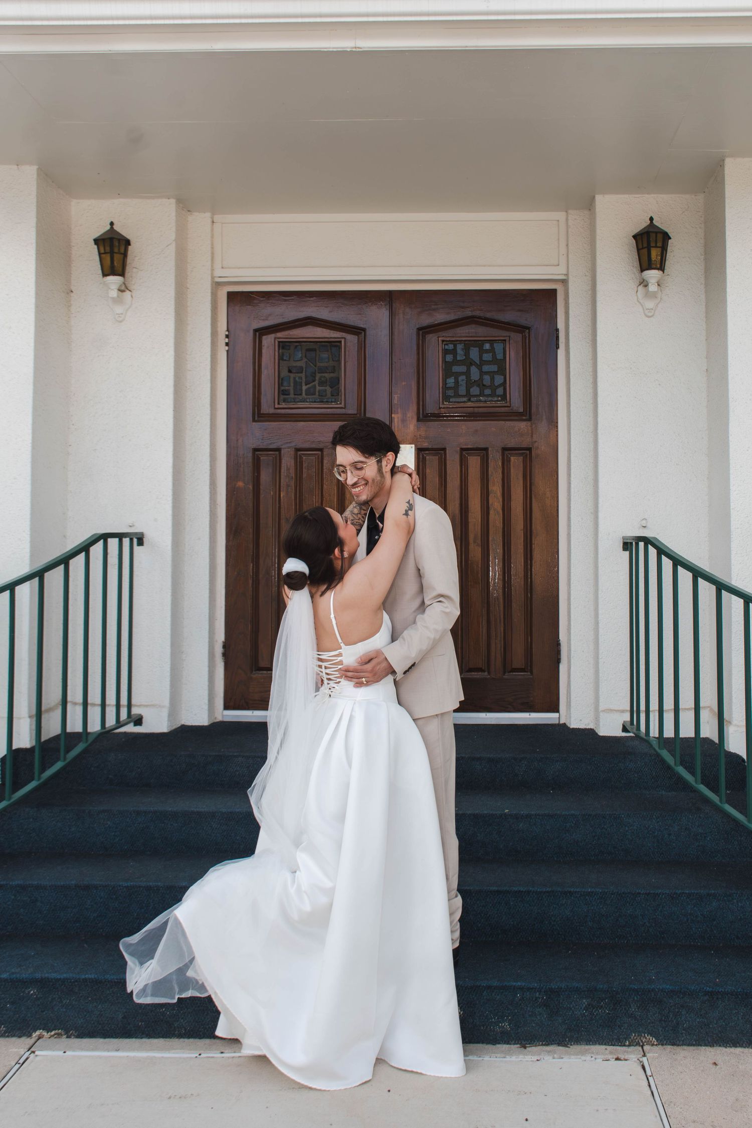 Bride and groom embrace and smile on steps before large wooden doors of a white building on wedding day.