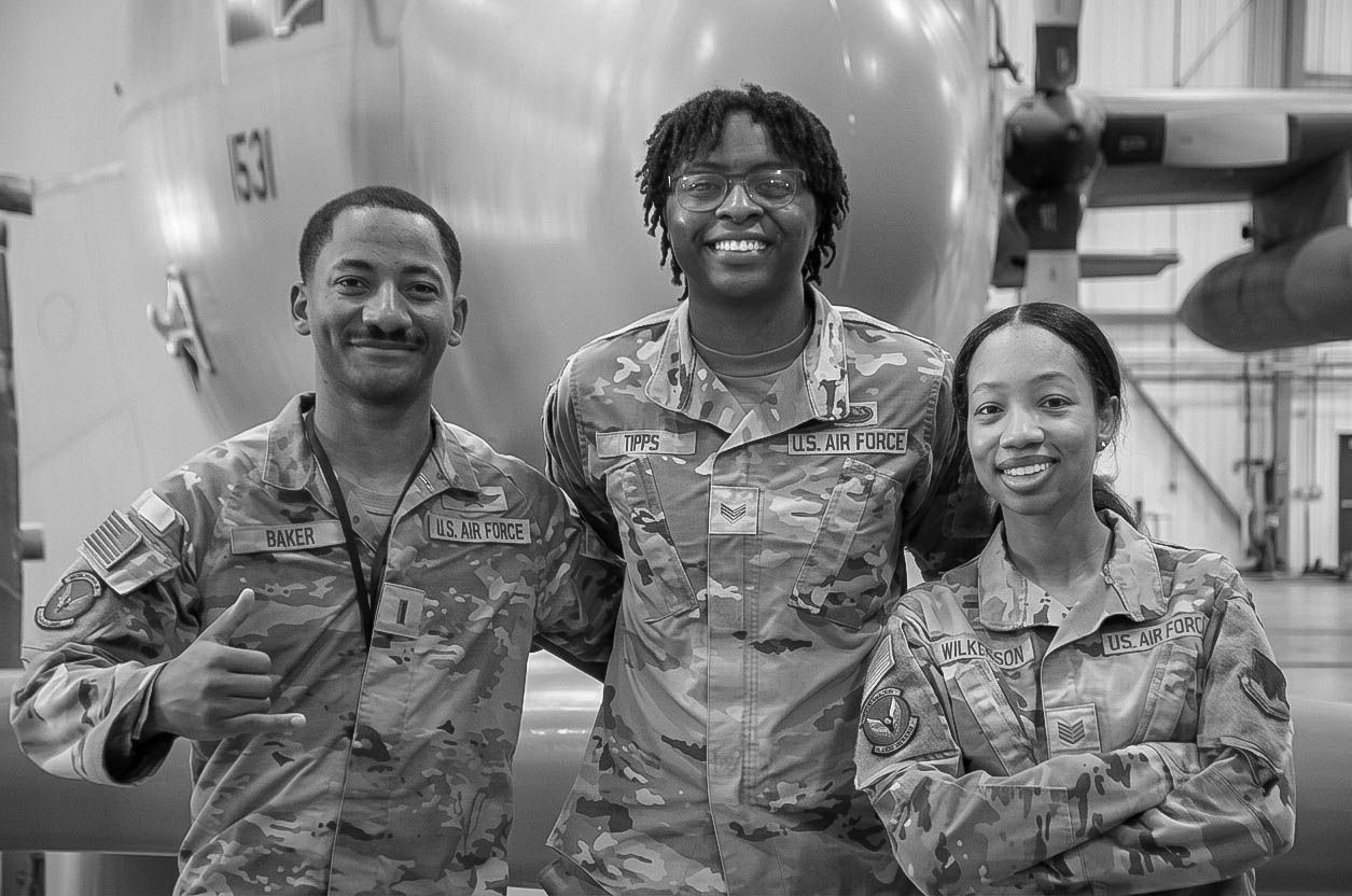 Three US Air Force airmen in camouflage uniforms smile together in a black and white photo.
