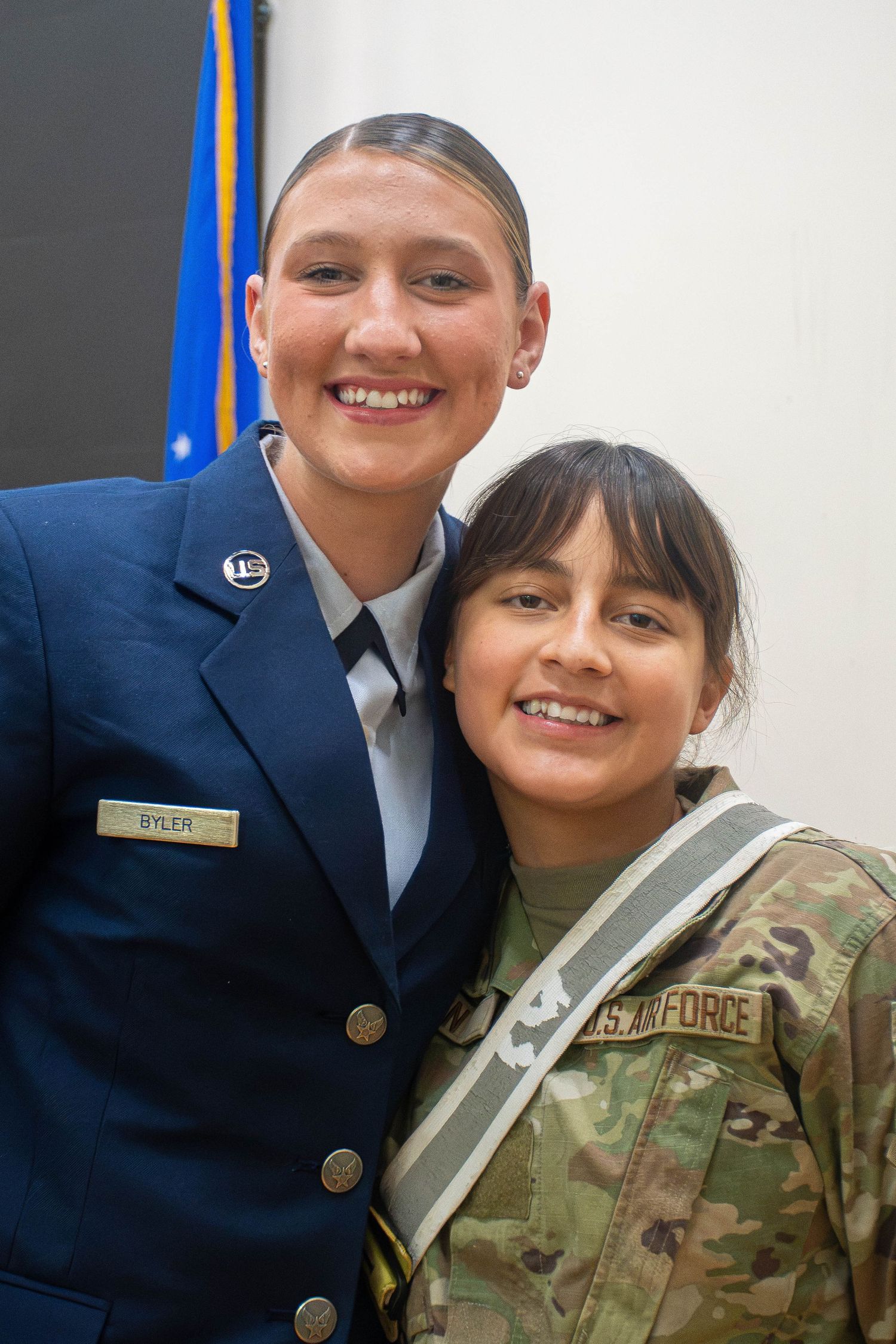 Two smiling US Air Force women, one in dress blues named Byler, posing together with American flag.