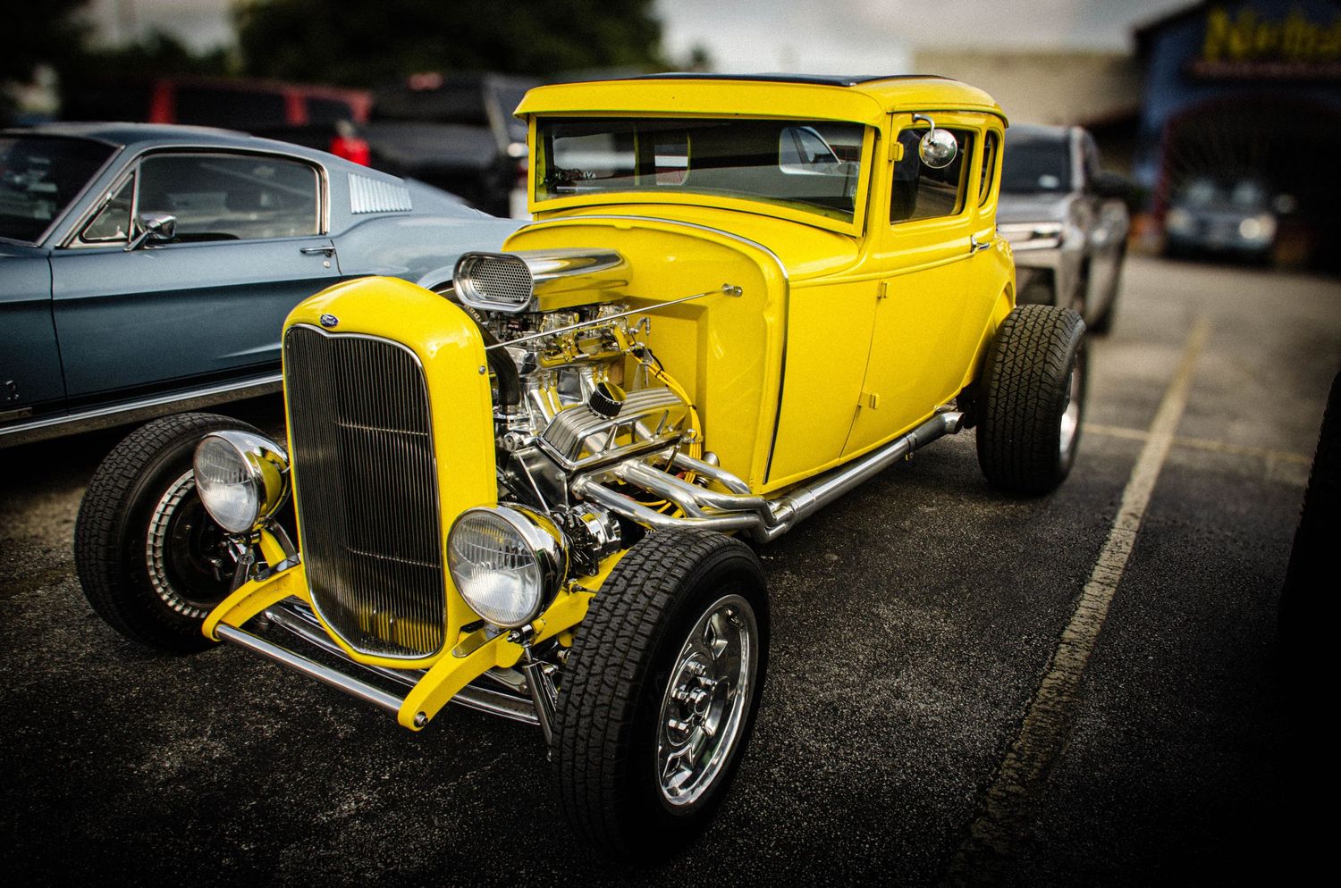Bright yellow vintage hot rod with exposed chrome engine on display at a classic car show in a parking lot.