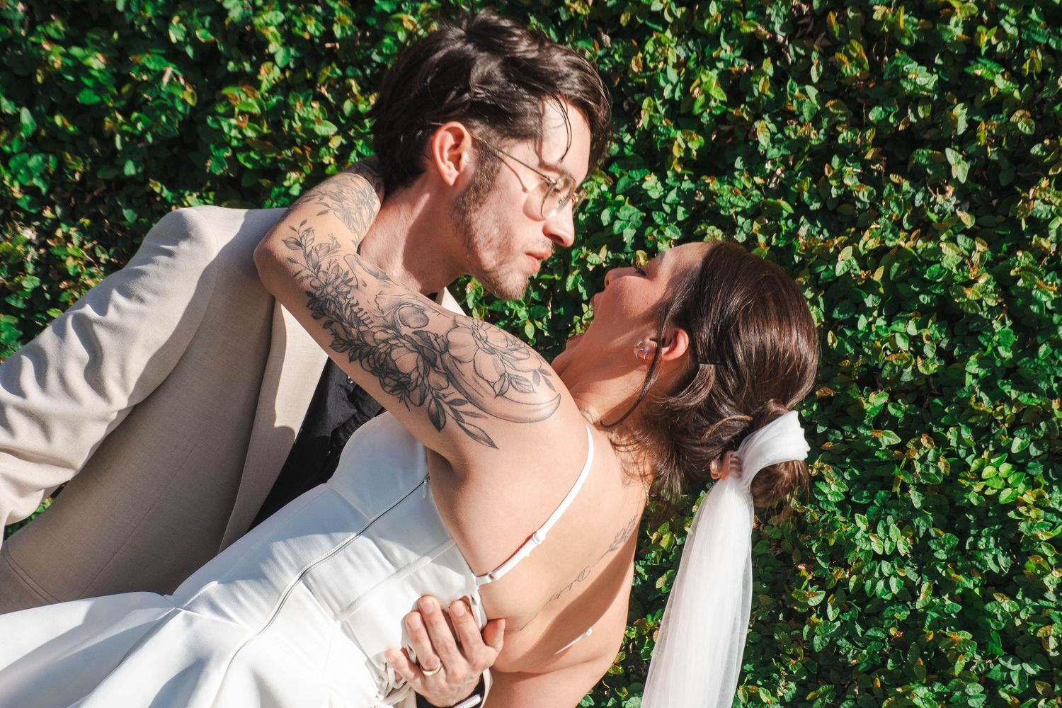 Couple sharing a romantic kiss on their wedding day in front of a lush green ivy wall.
