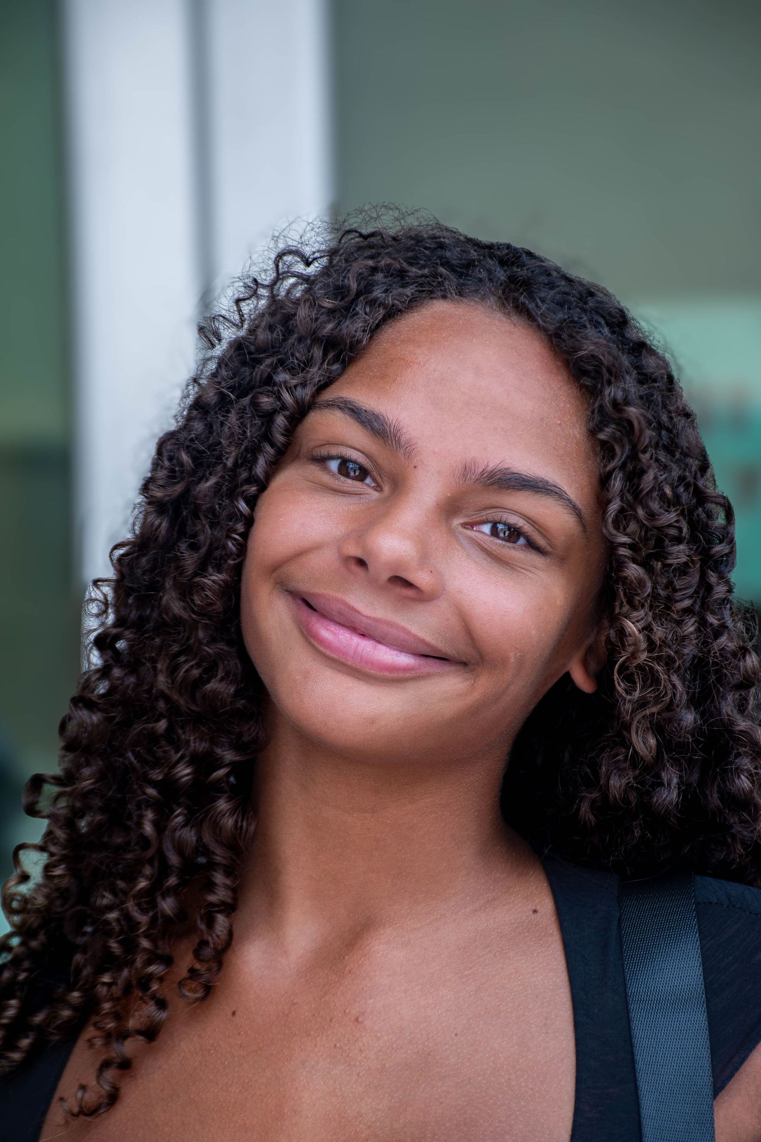 Young woman with long curly hair smiles warmly at camera outdoors.