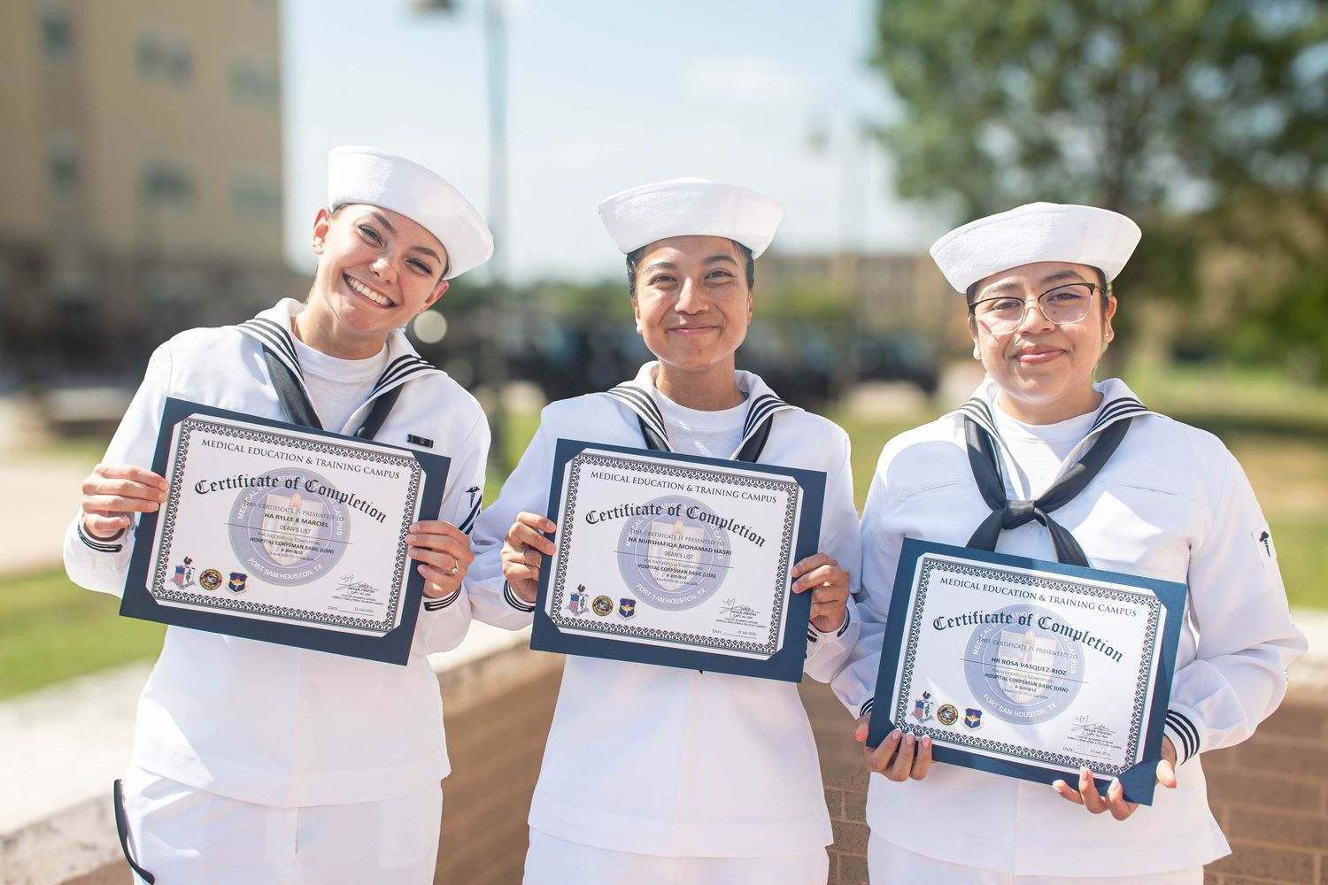 Three female Navy sailors smile holding Certificates of Completion at outdoor military graduation.