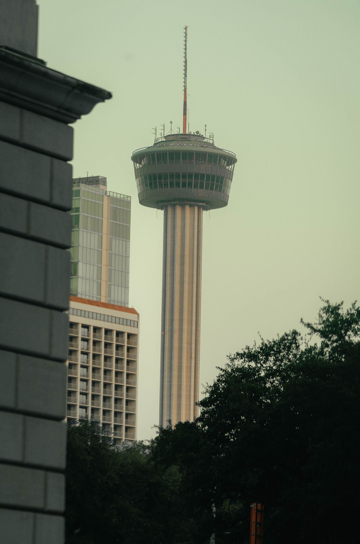 Tower of the Americas rises above San Antonio skyline surrounded by trees and buildings on a hazy day.