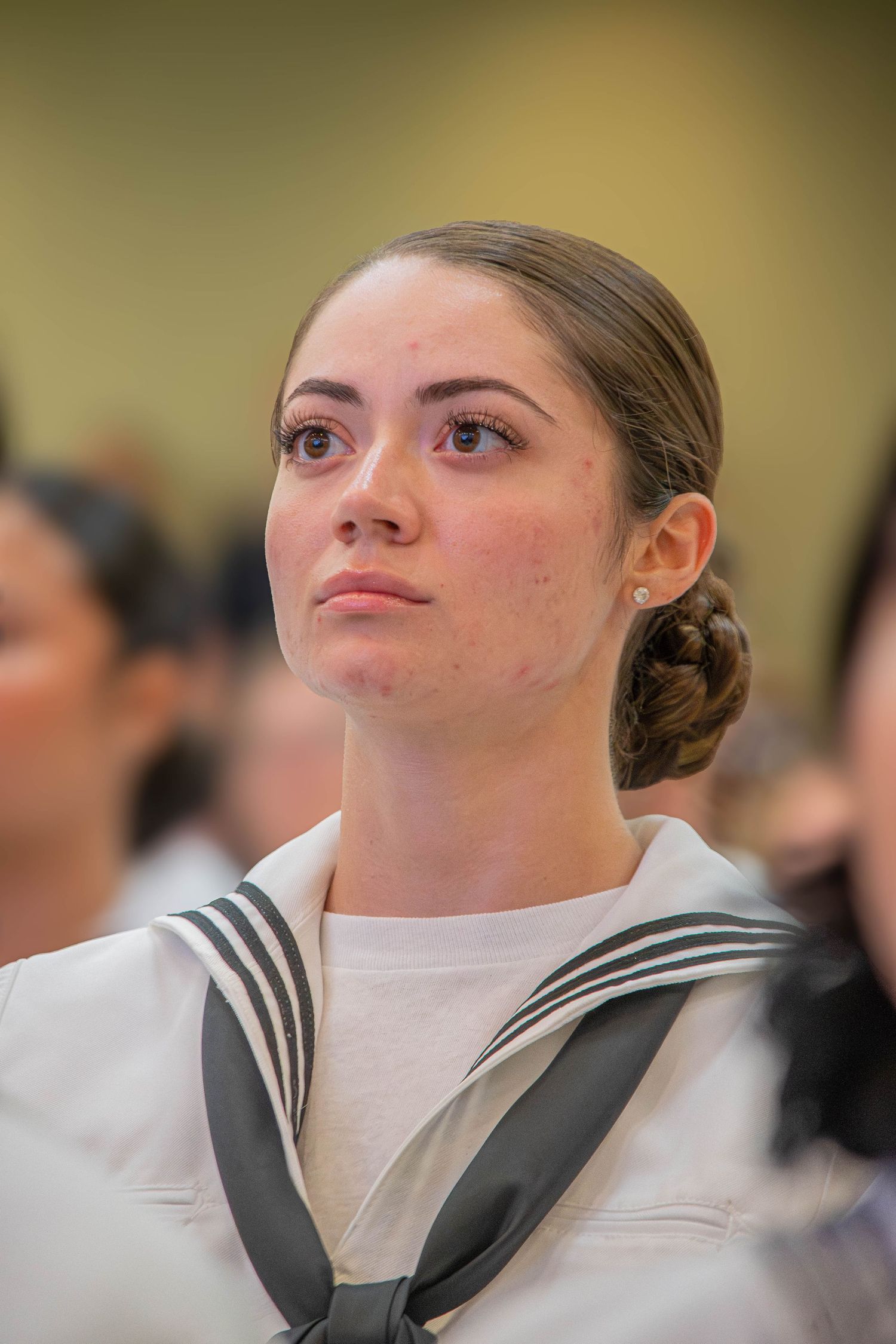 Young female Navy sailor with braided bun in white dress uniform looking upward attentively.
