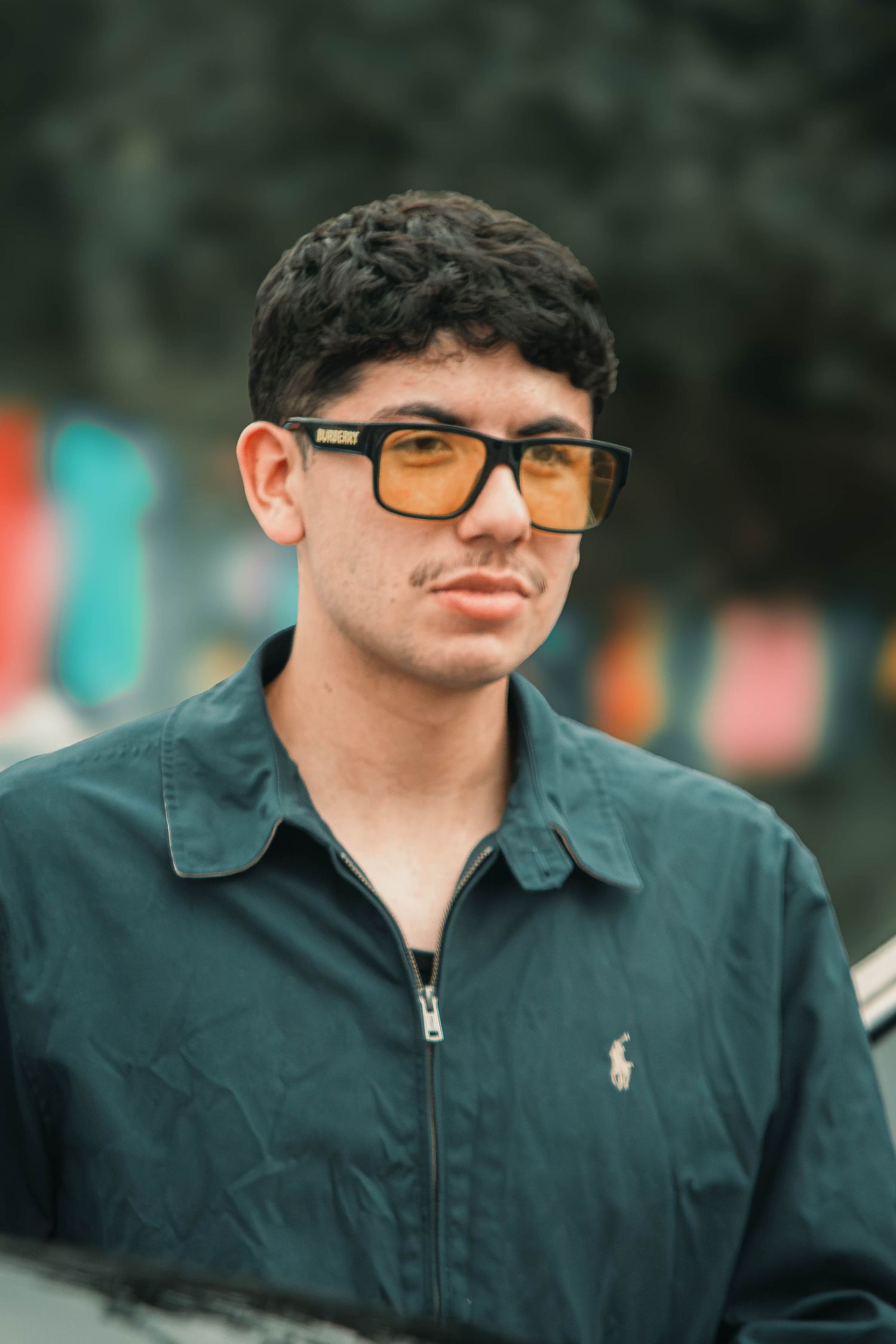 Young man with curly hair wearing orange-tinted square sunglasses and a teal Polo Ralph Lauren jacket.