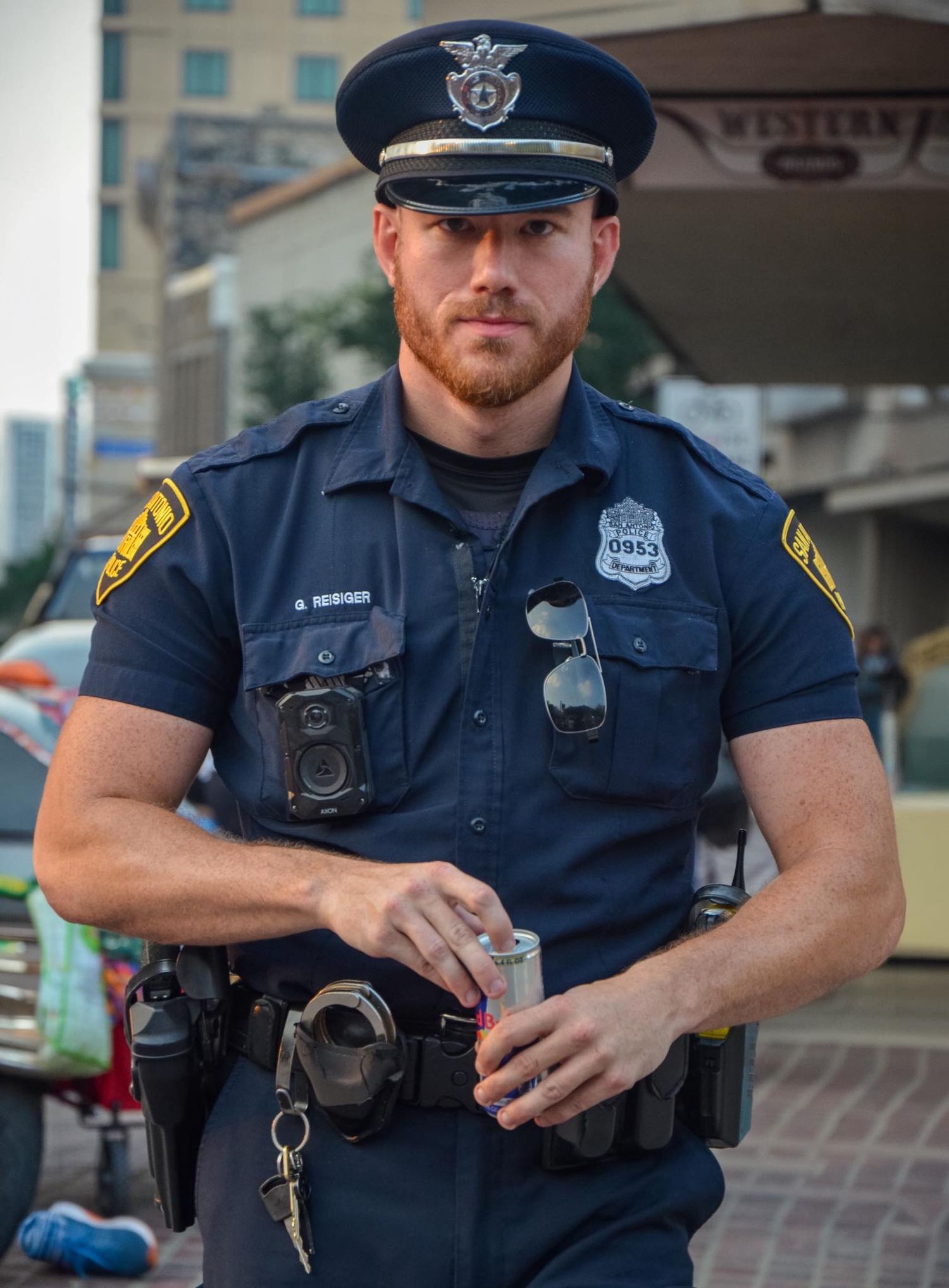 Muscular male police officer in uniform holding a canister outdoors in an urban setting.