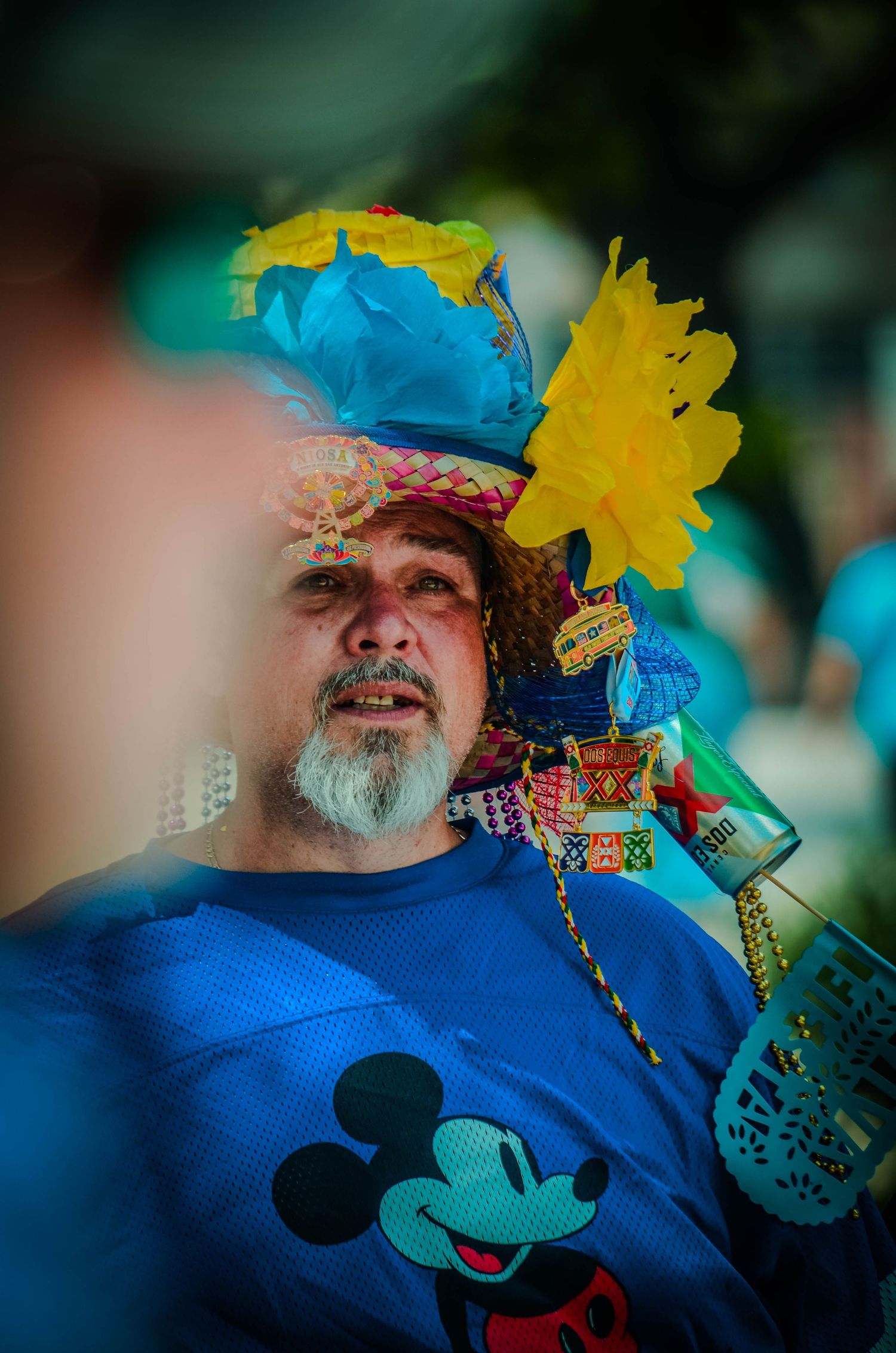 Elderly man wearing a colorful decorated hat and Mickey Mouse shirt at a festive outdoor event.