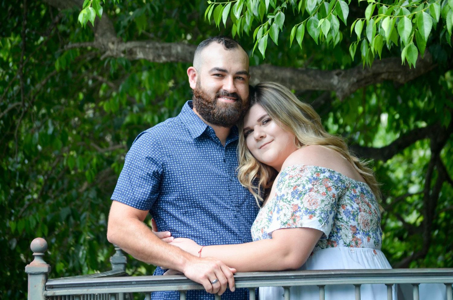 Happy couple poses on a bridge surrounded by lush green trees in an outdoor engagement photo session.