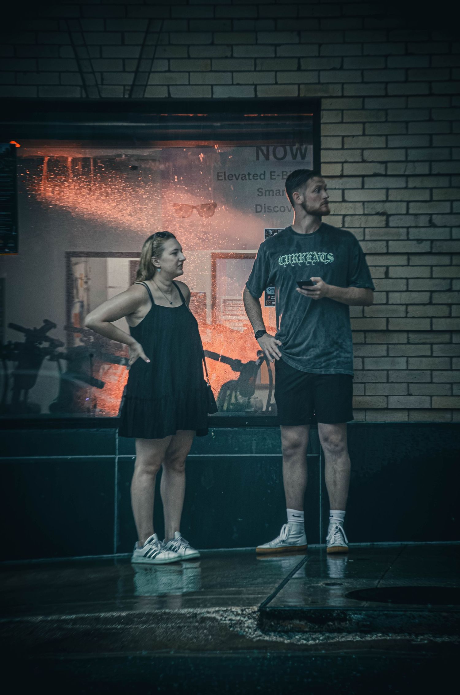 A man and woman stand outside a building on a rainy night, sheltering under an awning on wet pavement.