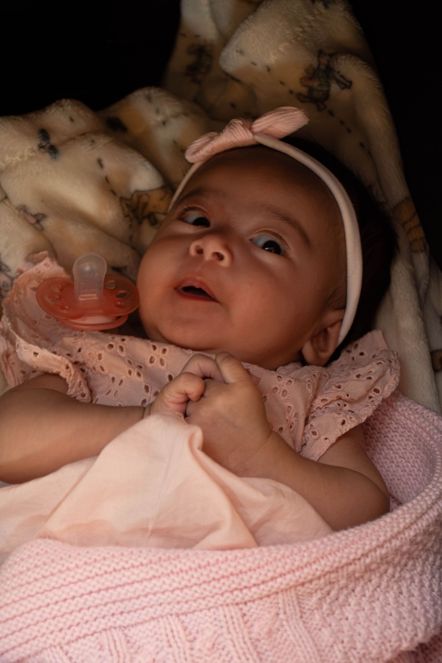 Adorable baby girl in pink outfit with bow headband and pacifier lying on blanket.
