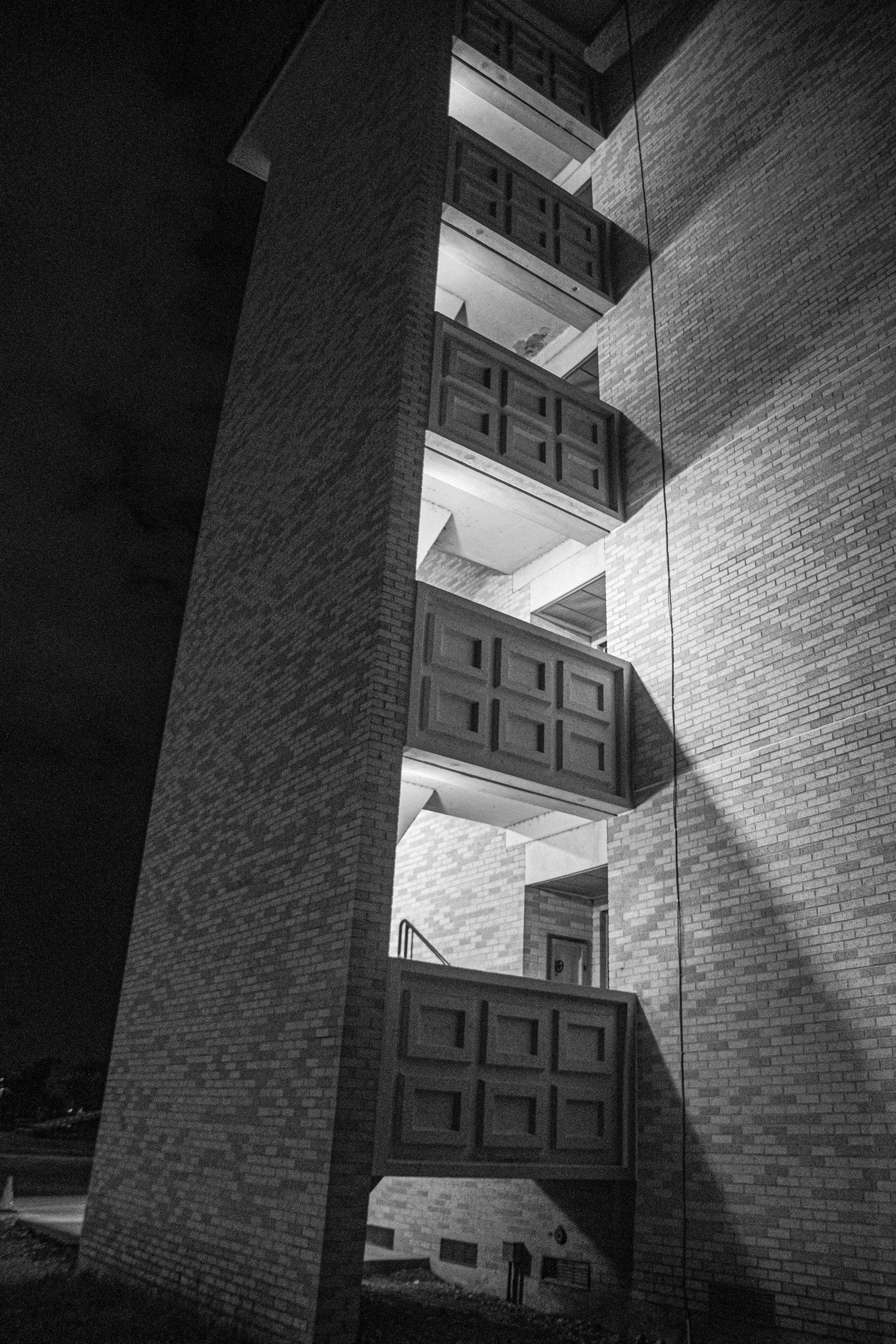Low angle black and white view of a brick building with stacked cantilevered balconies lit at night.