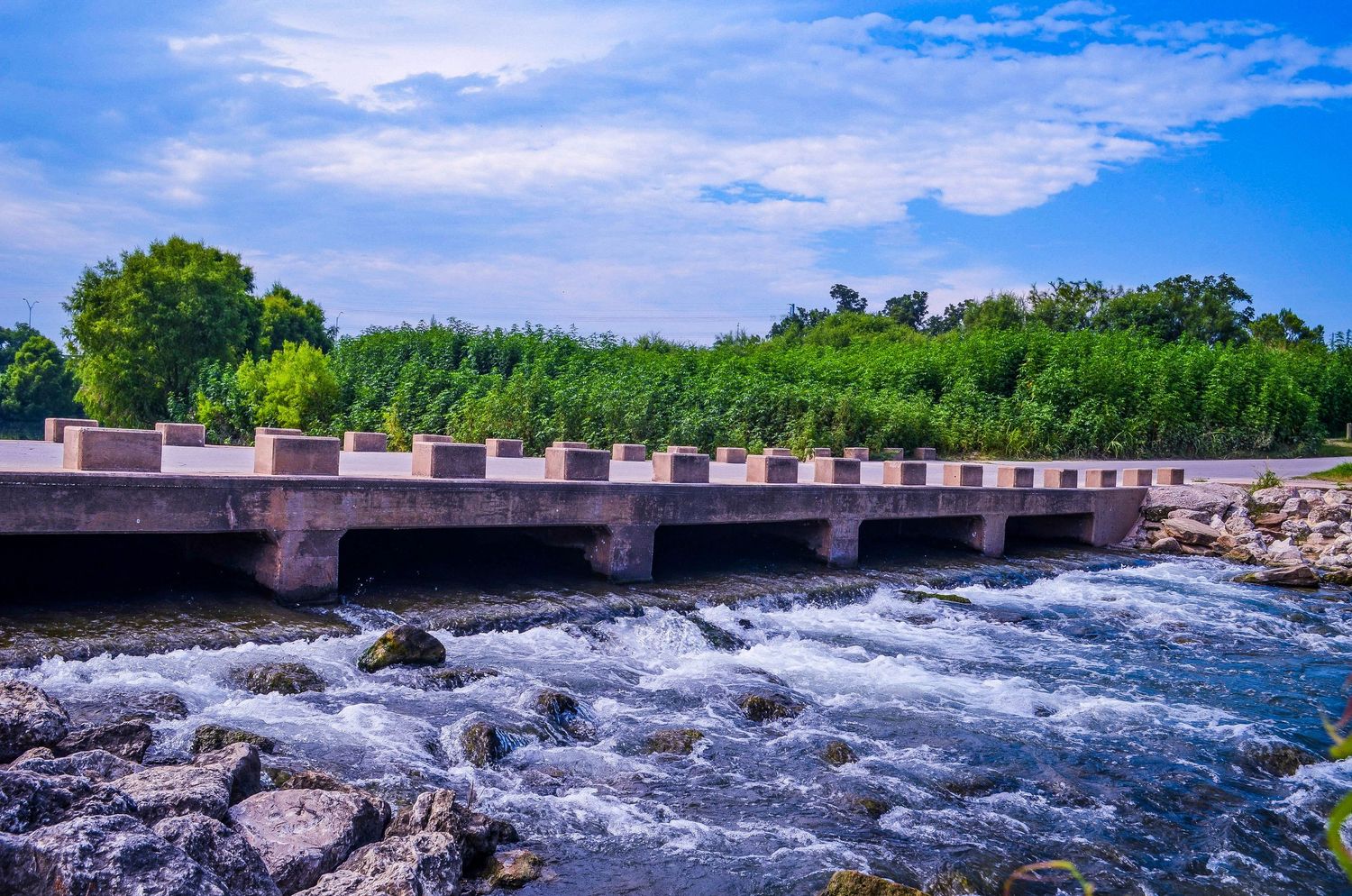 A concrete dam with rushing white water rapids flowing over rocks under a blue sky.