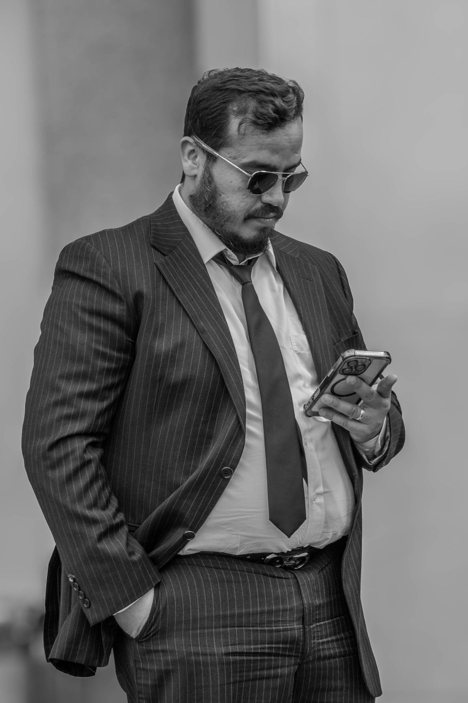 Black and white photo of a bearded man in a suit and sunglasses using a smartphone.