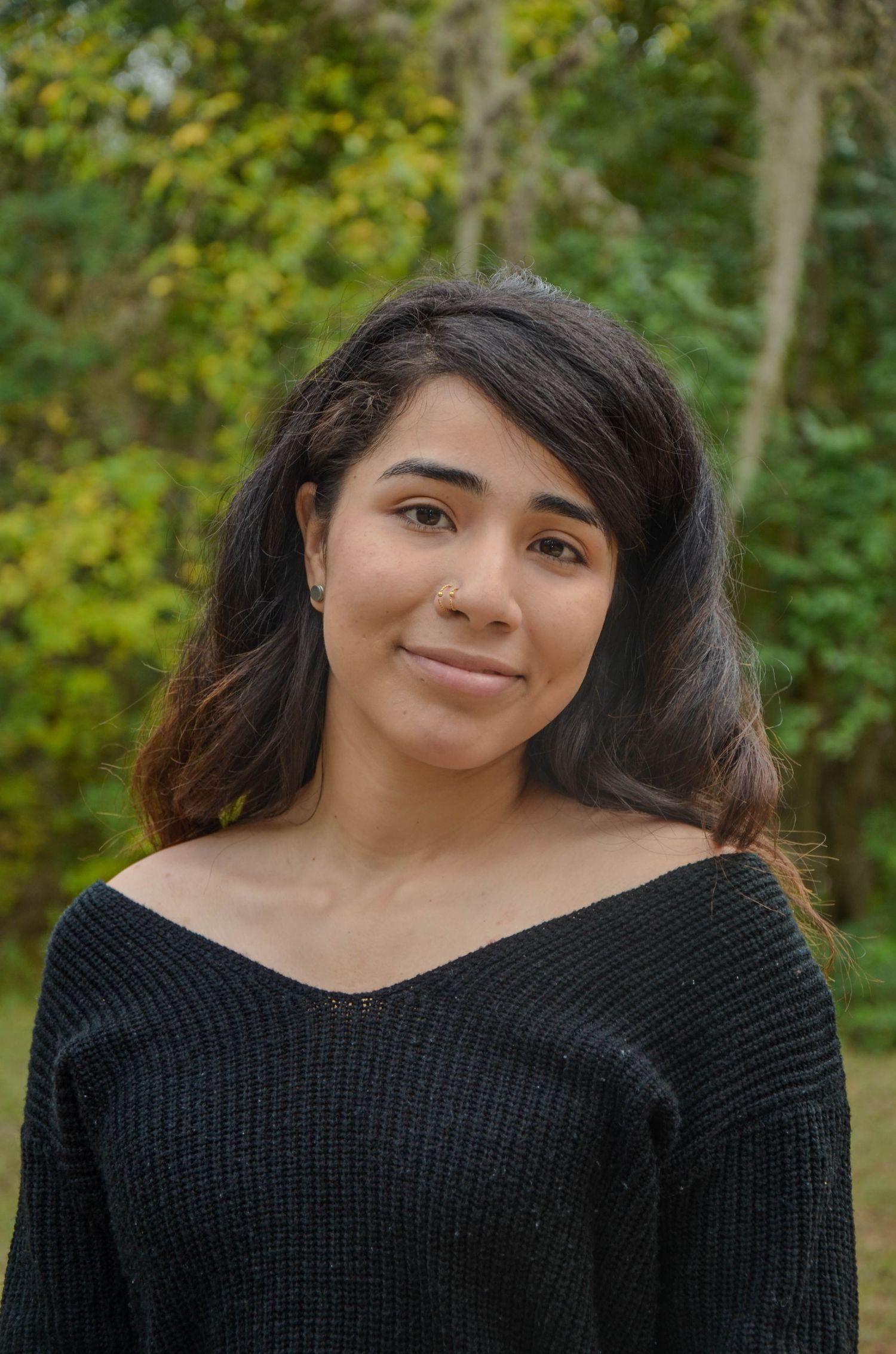 Young woman with dark hair smiling outdoors wearing black off-shoulder sweater with green trees in background.