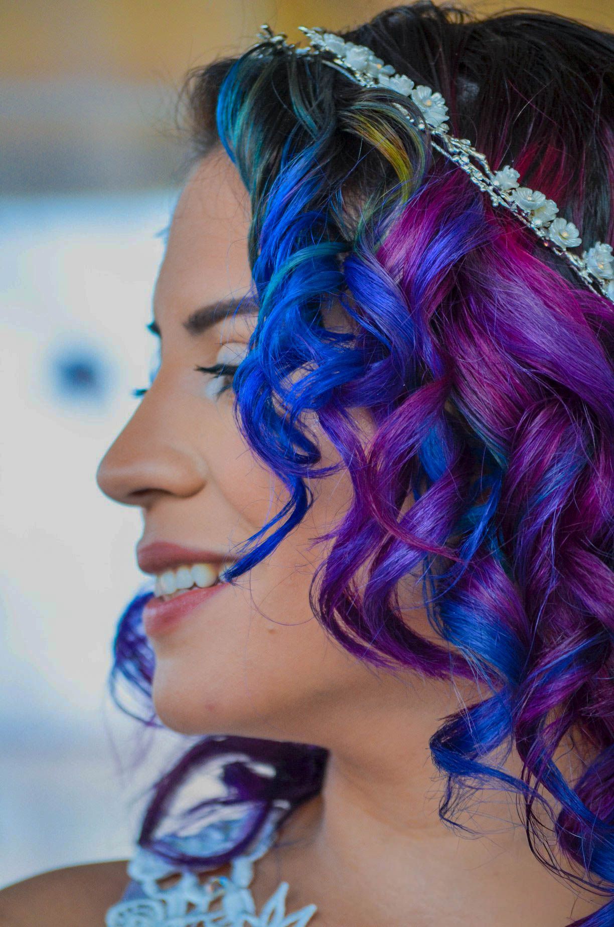 A close-up profile of a woman with vibrant blue and purple curly hair wearing a floral headband.