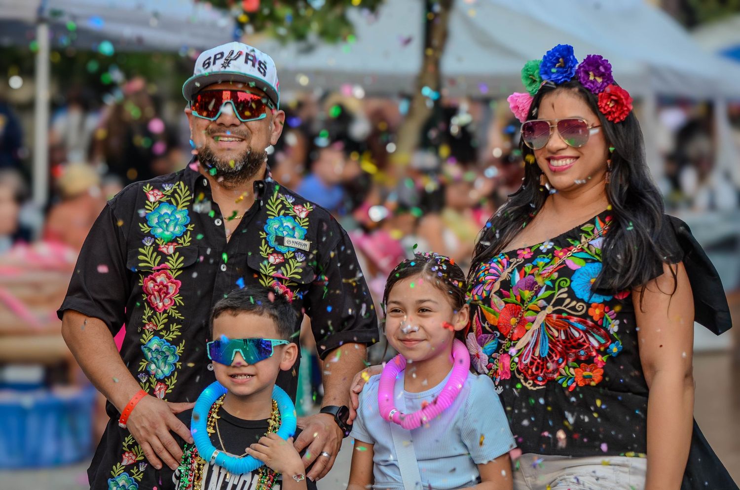 Family of four in matching black embroidered outfits celebrating at colorful outdoor festival.