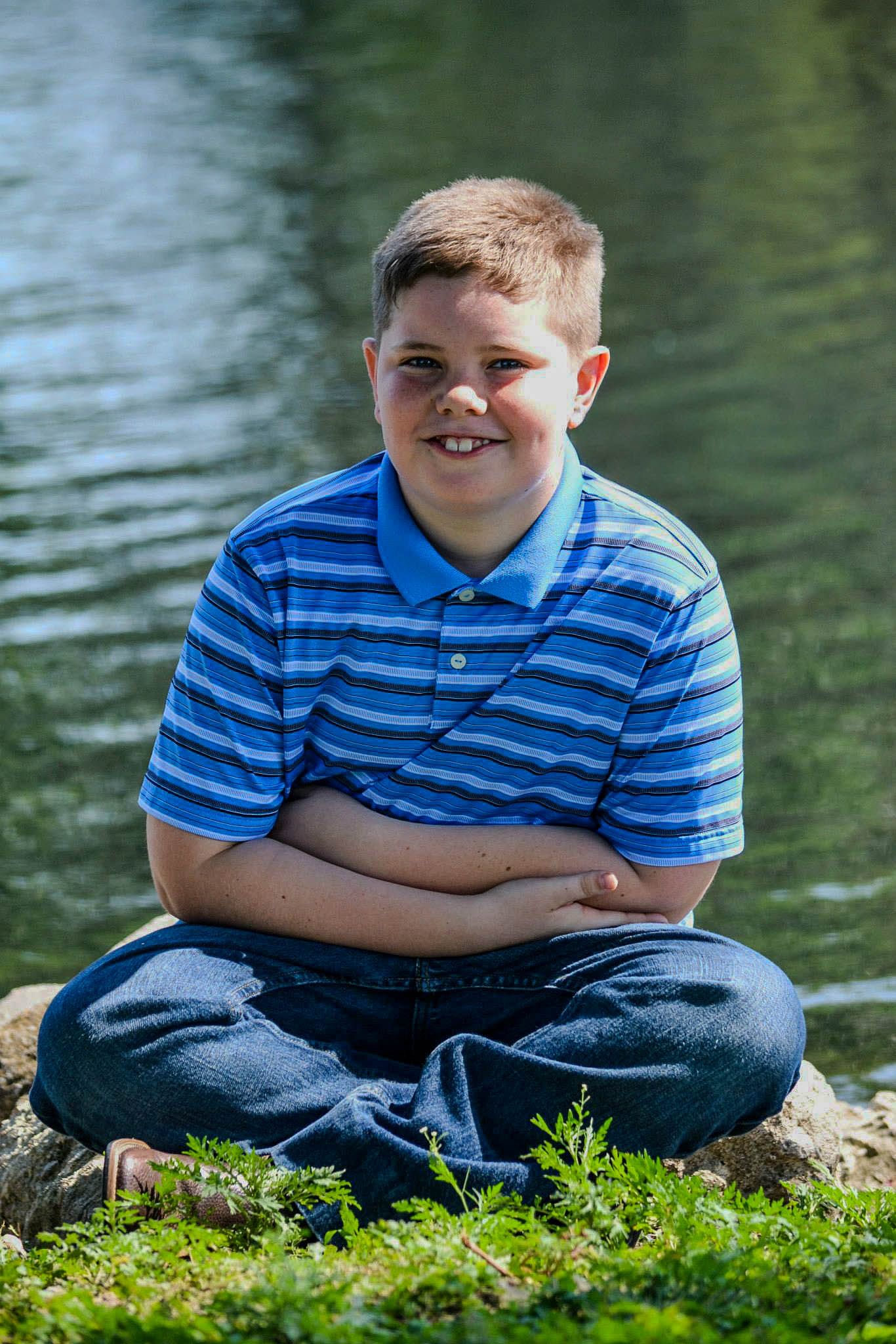 Smiling boy in blue striped polo sitting cross-legged by a pond.