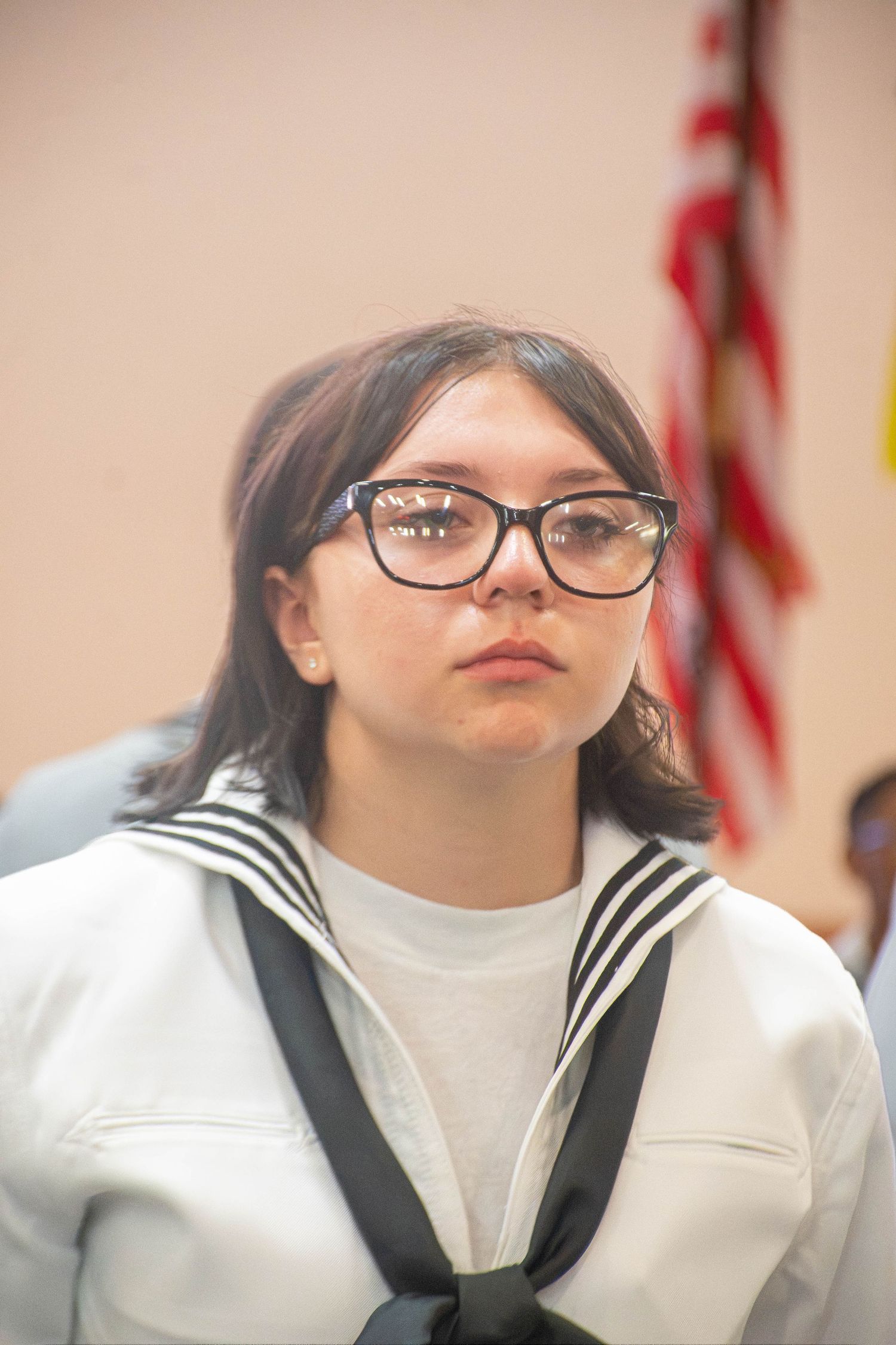 Young female Navy recruit wearing glasses in white sailor uniform stands at attention during graduation.
