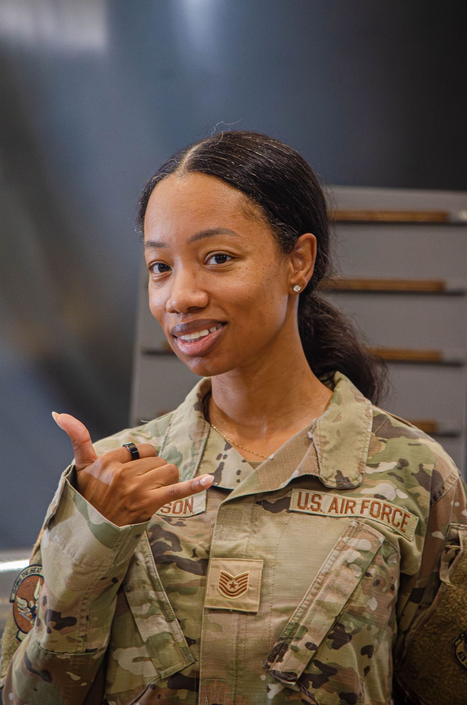 A female US Air Force Tech Sergeant in camouflage uniform makes a shaka hand gesture.