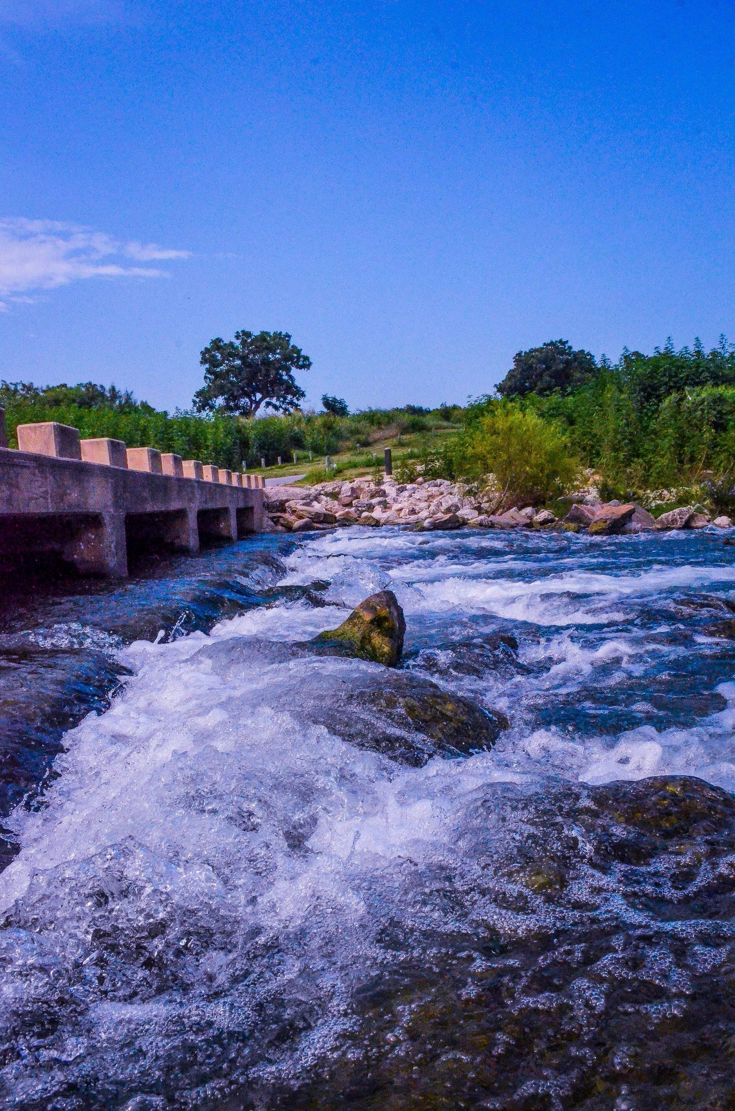 Rushing white water rapids over rocks beside a concrete bridge under a bright blue sky.