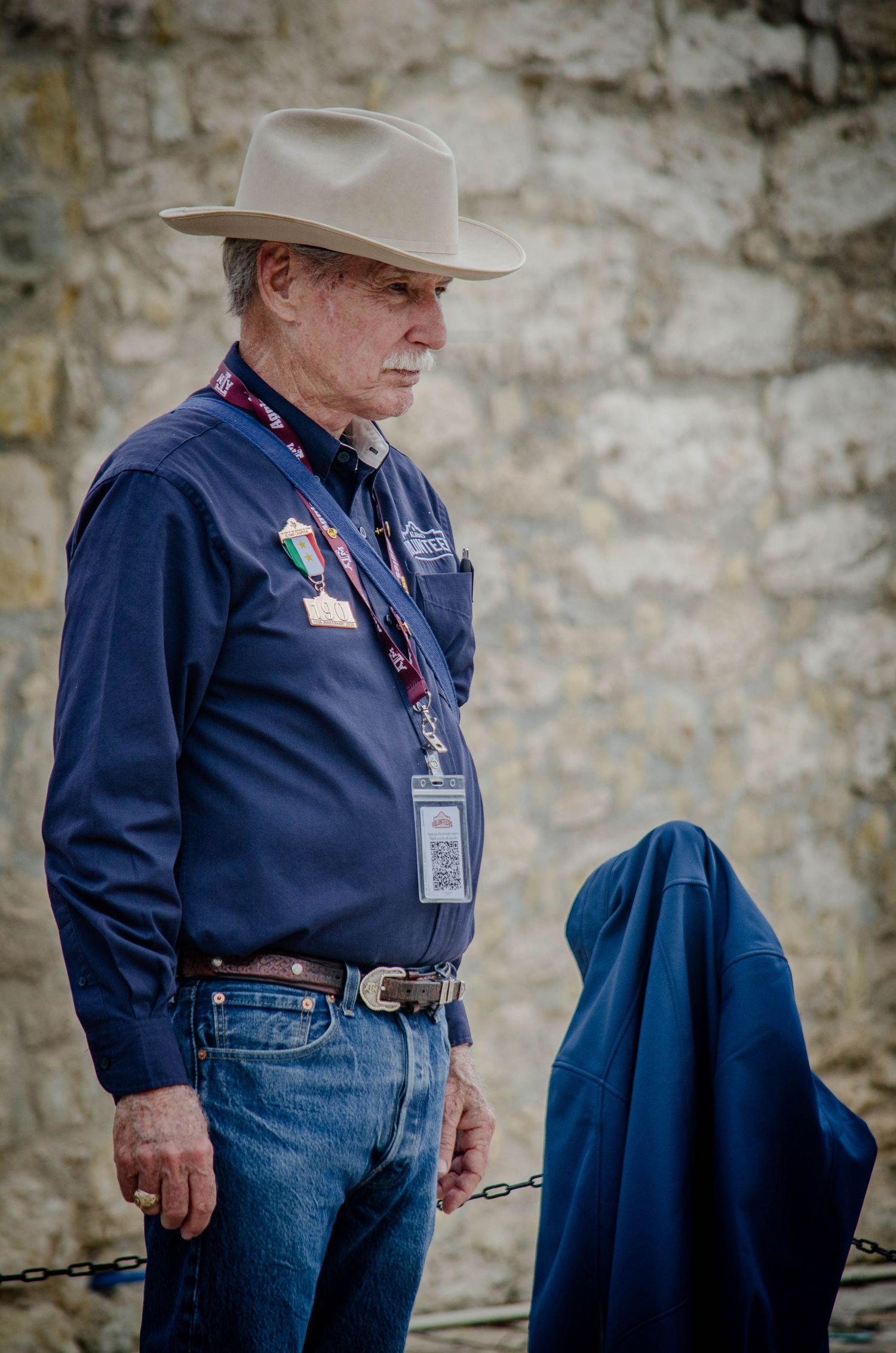 Senior man in cowboy hat and blue shirt with ID badge standing near stone wall outdoors.