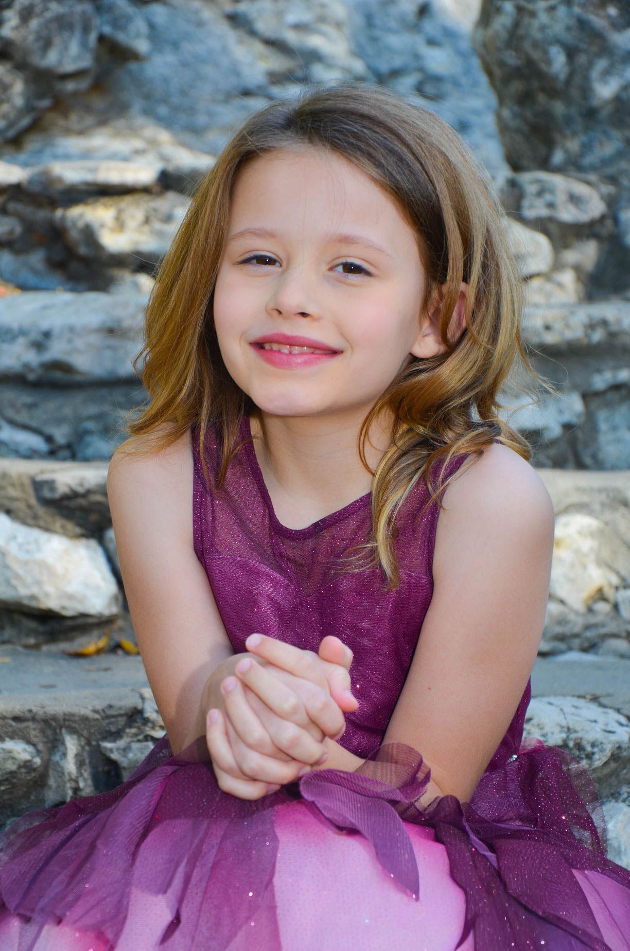 Cute young girl in a purple tutu dress smiles with clasped hands while sitting on stone steps.