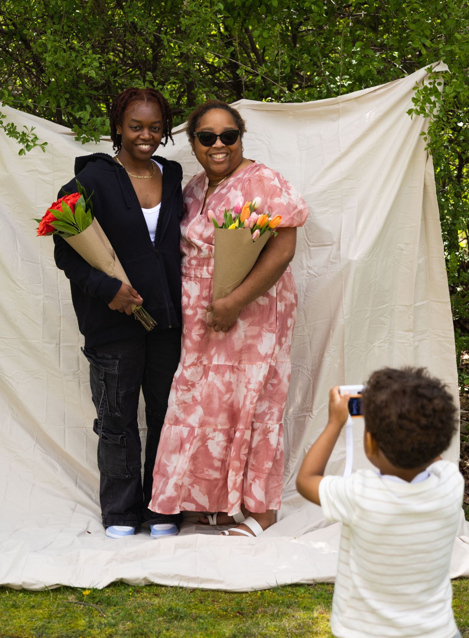 Portrait of two women posing outdoors holding flower bouquets while a little boy takes their photo