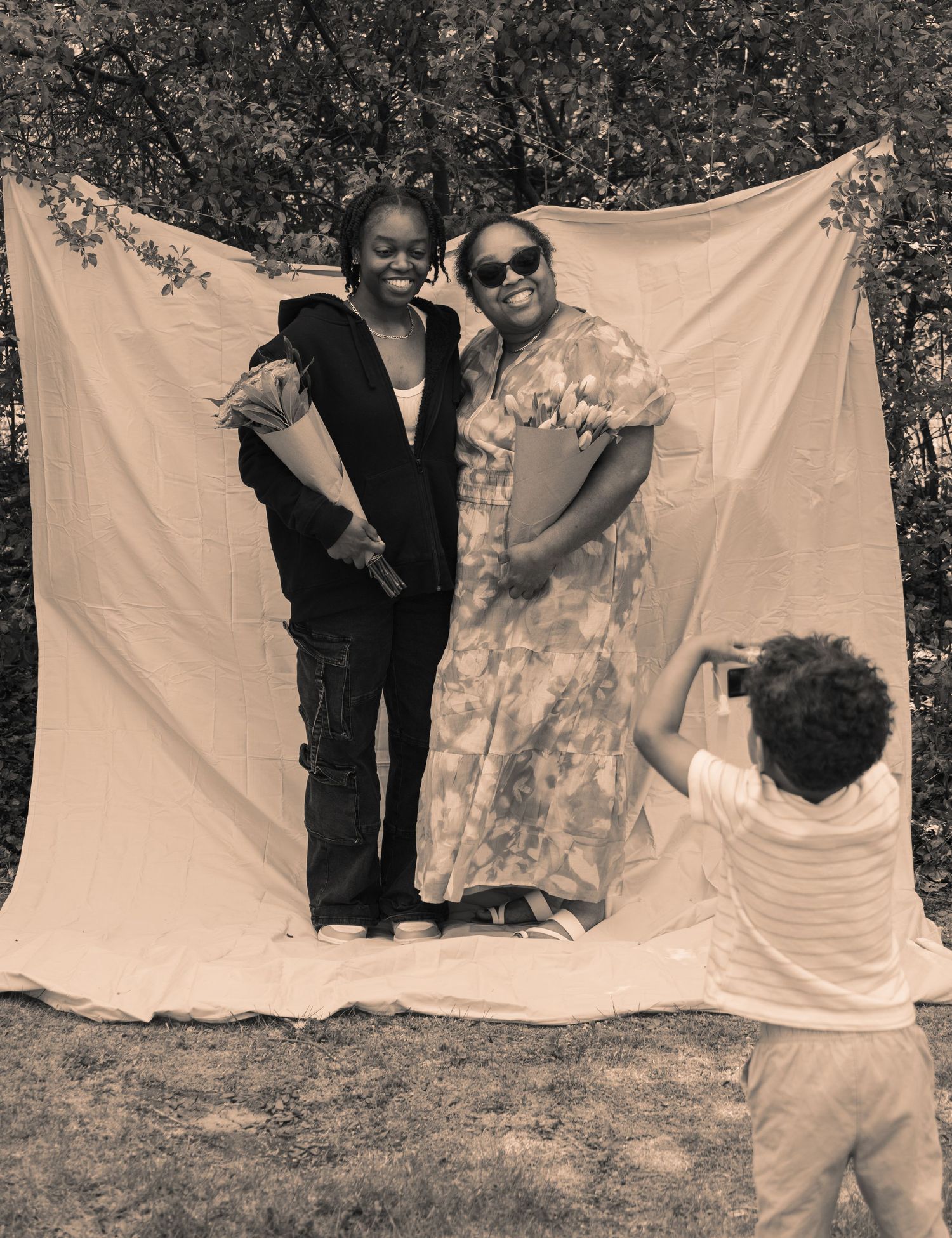 Black and white portrait of two women posing outdoors holding flower bouquets while a little boy takes their photo
