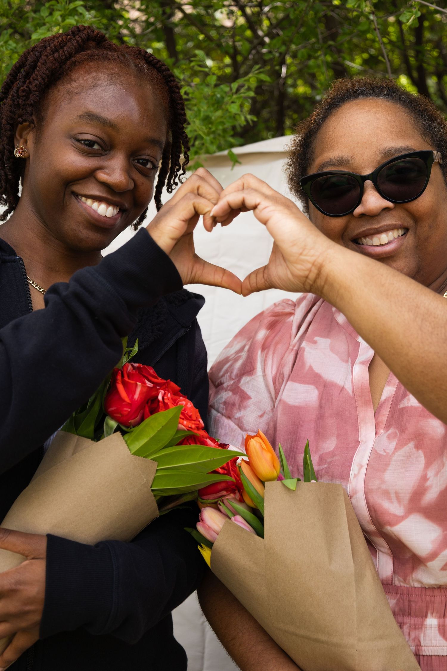 Two women posing outdoors holding flower bouquets, making a heart shape with their hands