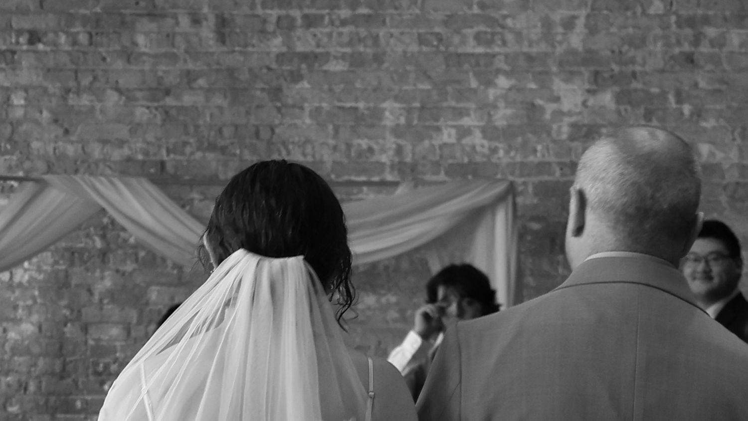 Black and white photo of a bride in veil standing at wedding altar with officiant and guests in brick venue.