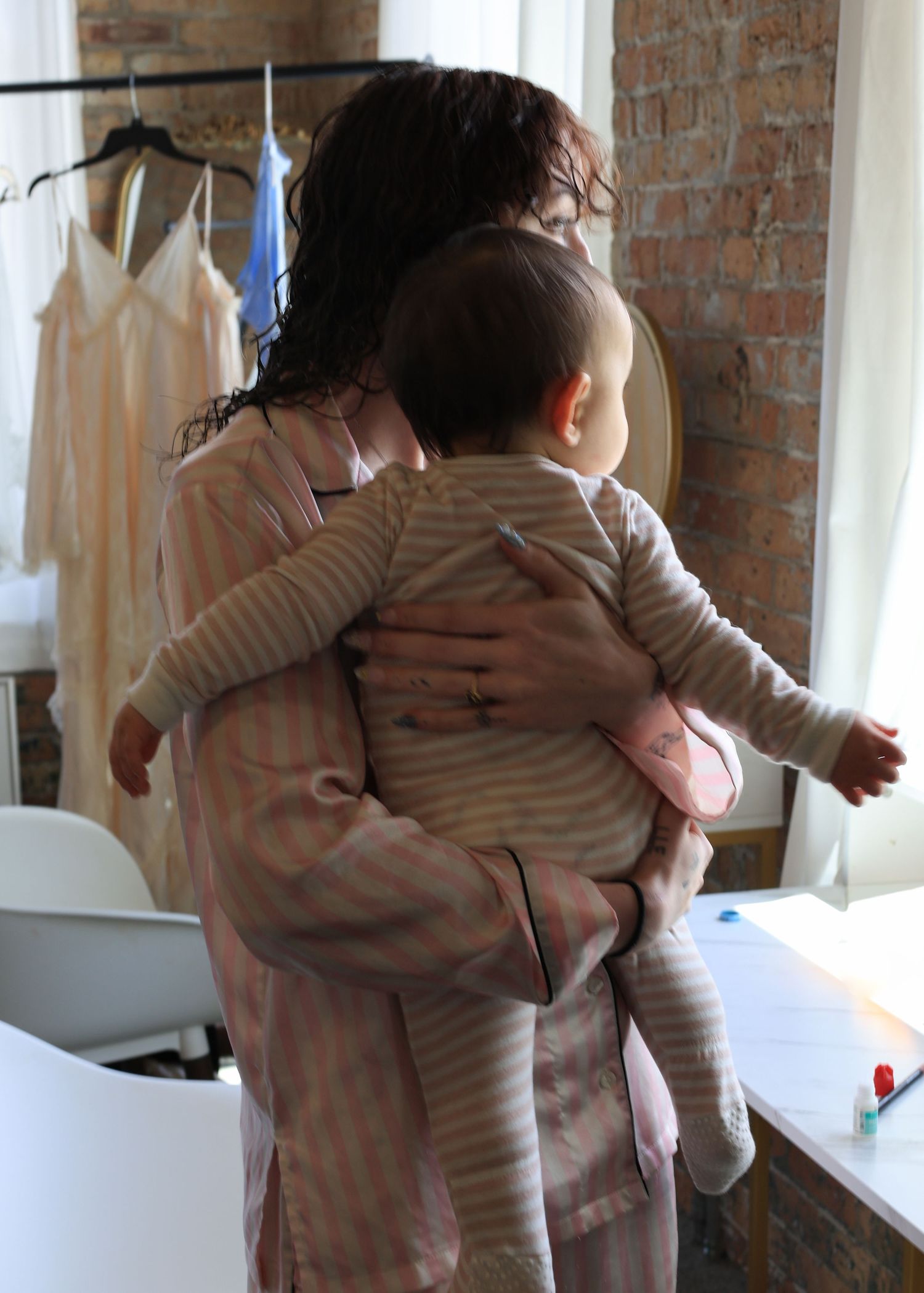 Mother holding baby infant in striped outfit near window in bright room with dresses.