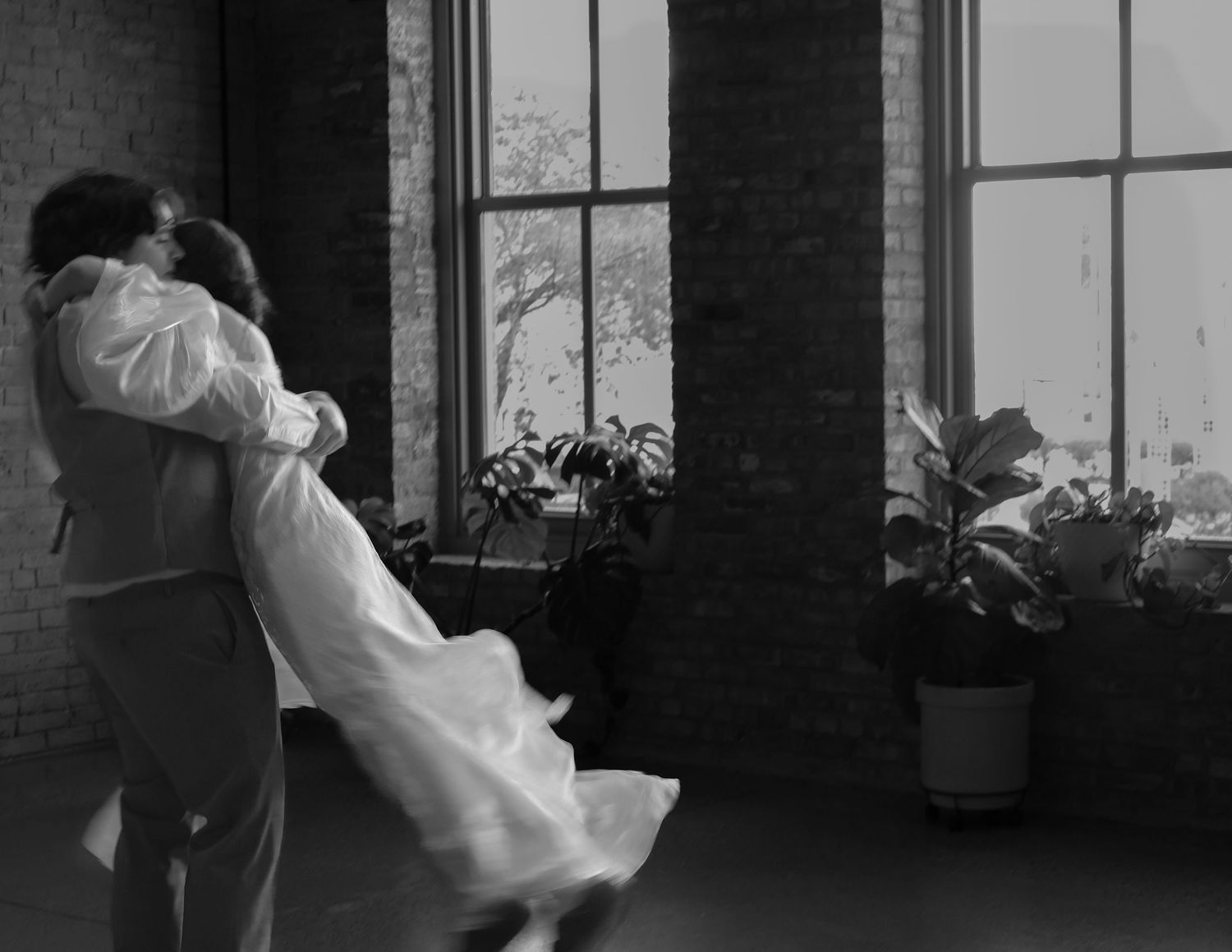 Black and white photo of couple dancing and embracing near large windows in brick venue.
