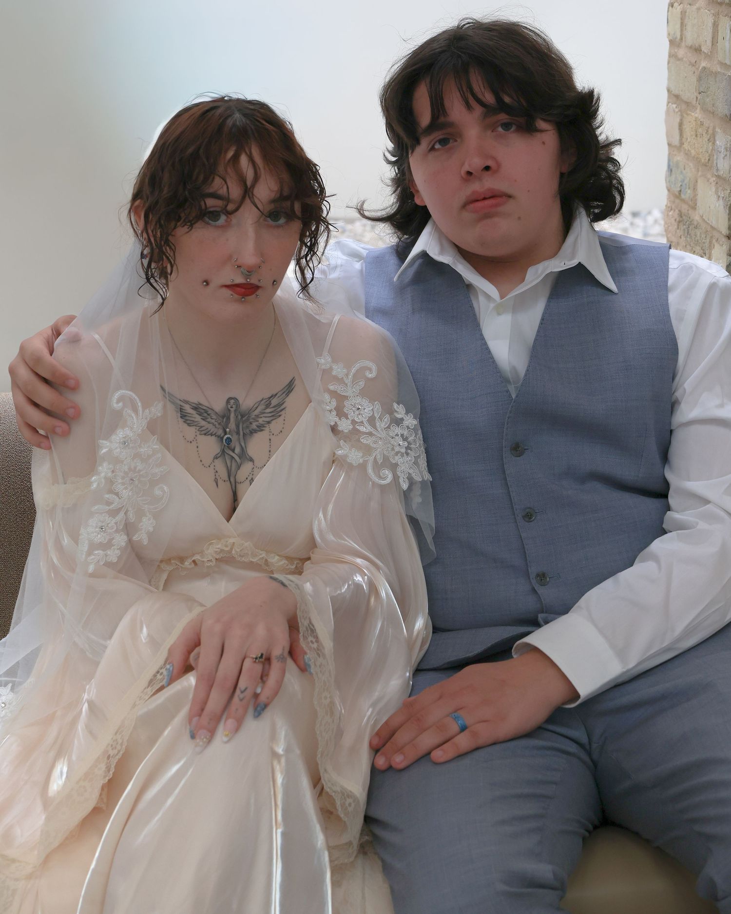 A couple in wedding attire, woman in lace gown with tattoos, man in gray vest, posing together indoors.