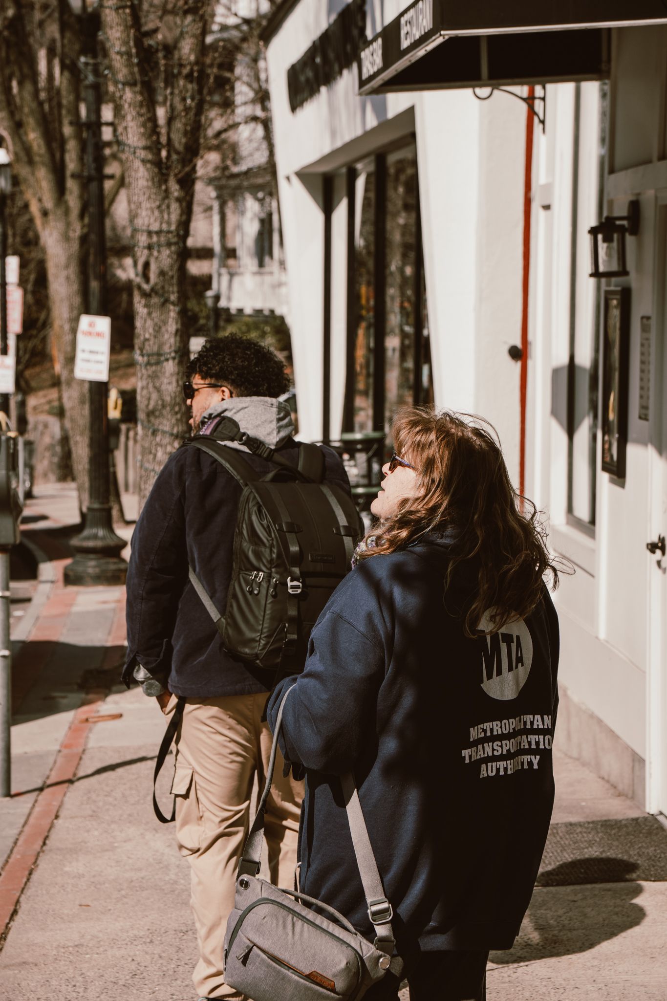 Two young people with backpacks walking on a sunny tree-lined urban sidewalk.
