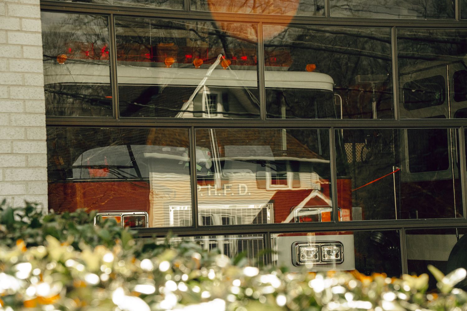 Close-up of fire truck marked H.F.D. visible through reflective glass firehouse garage doors with shrubs.