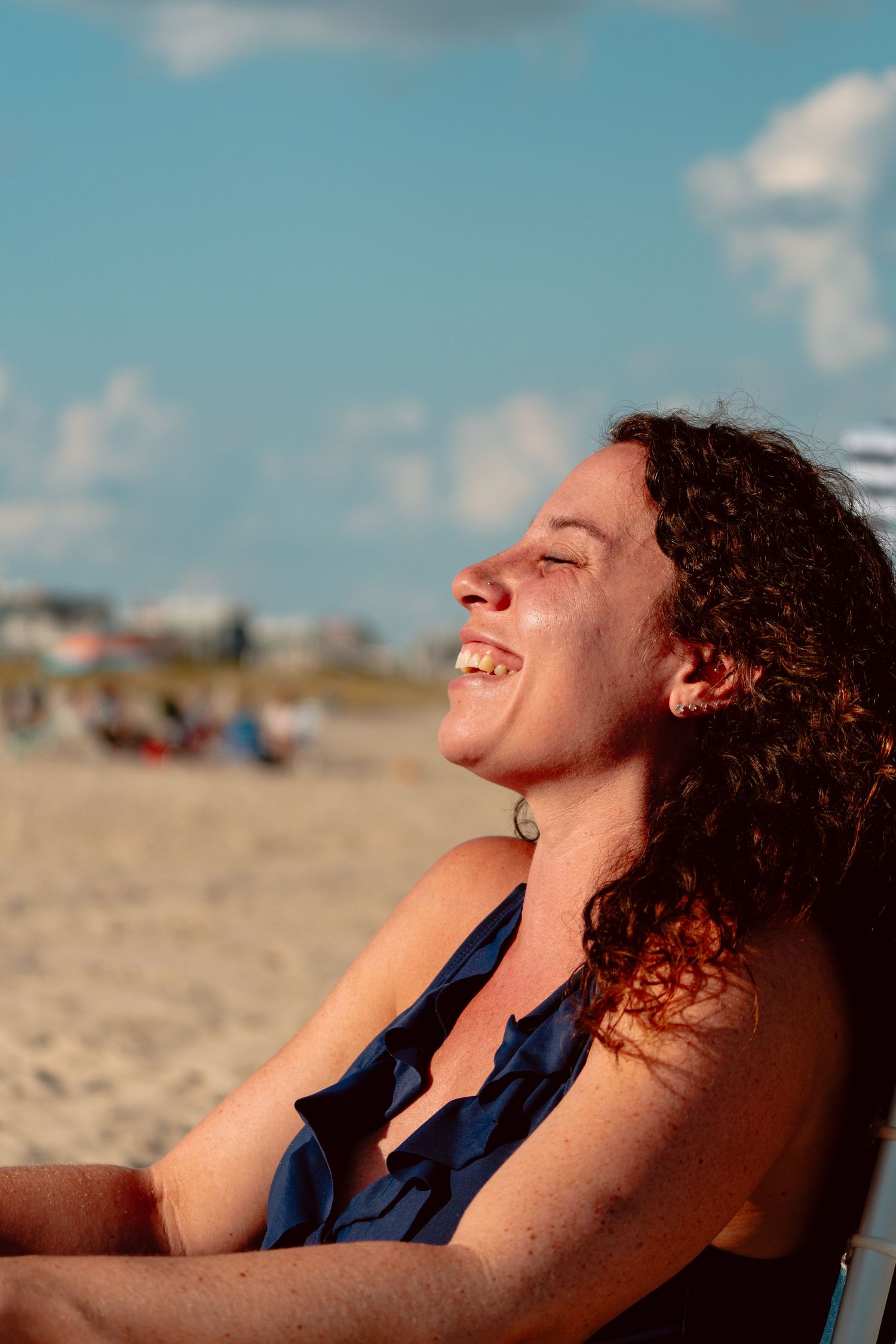 A curly-haired woman laughs joyfully while sunbathing on a sandy beach on a sunny day.