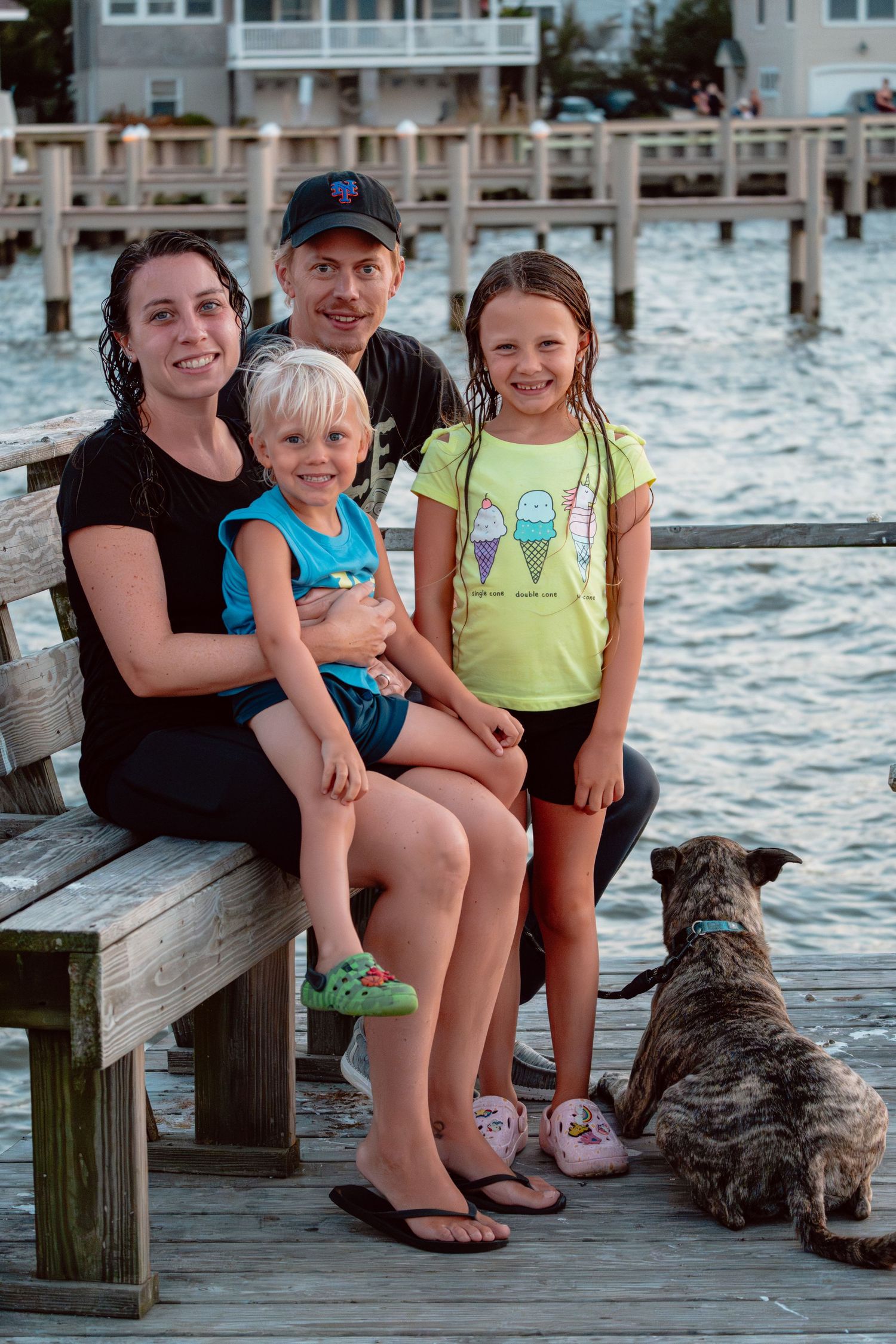 Happy family with two kids and a dog sitting on a wooden dock by the water at dusk.
