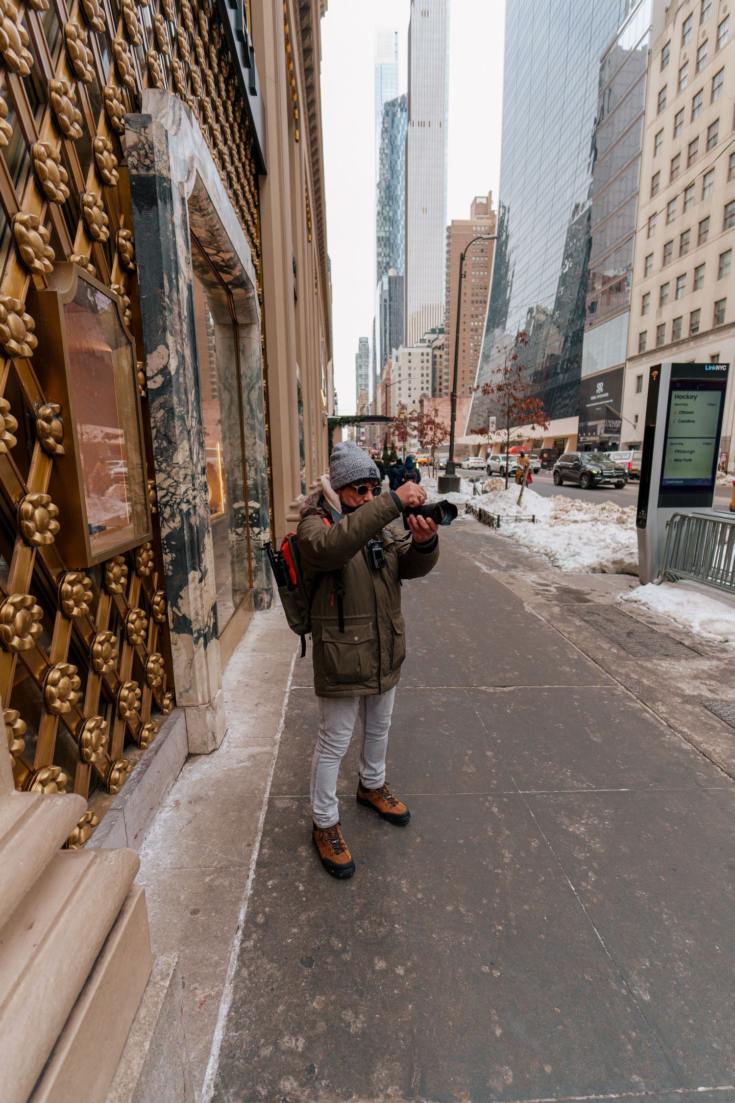 A photographer in a green parka photographs ornate golden doors on a snowy New York City sidewalk.