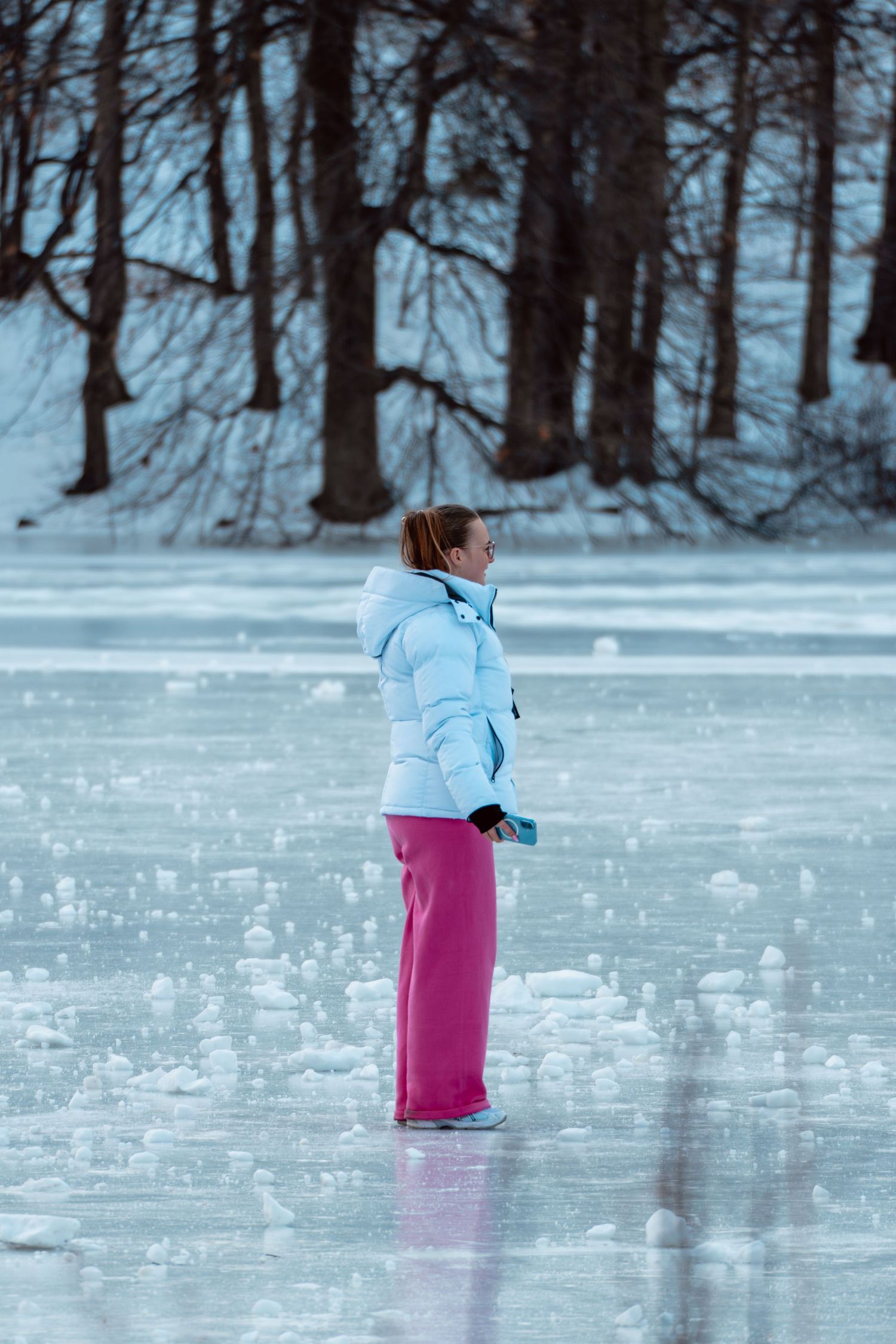 A young woman in a light blue jacket and pink pants ice skates on a frozen outdoor rink.